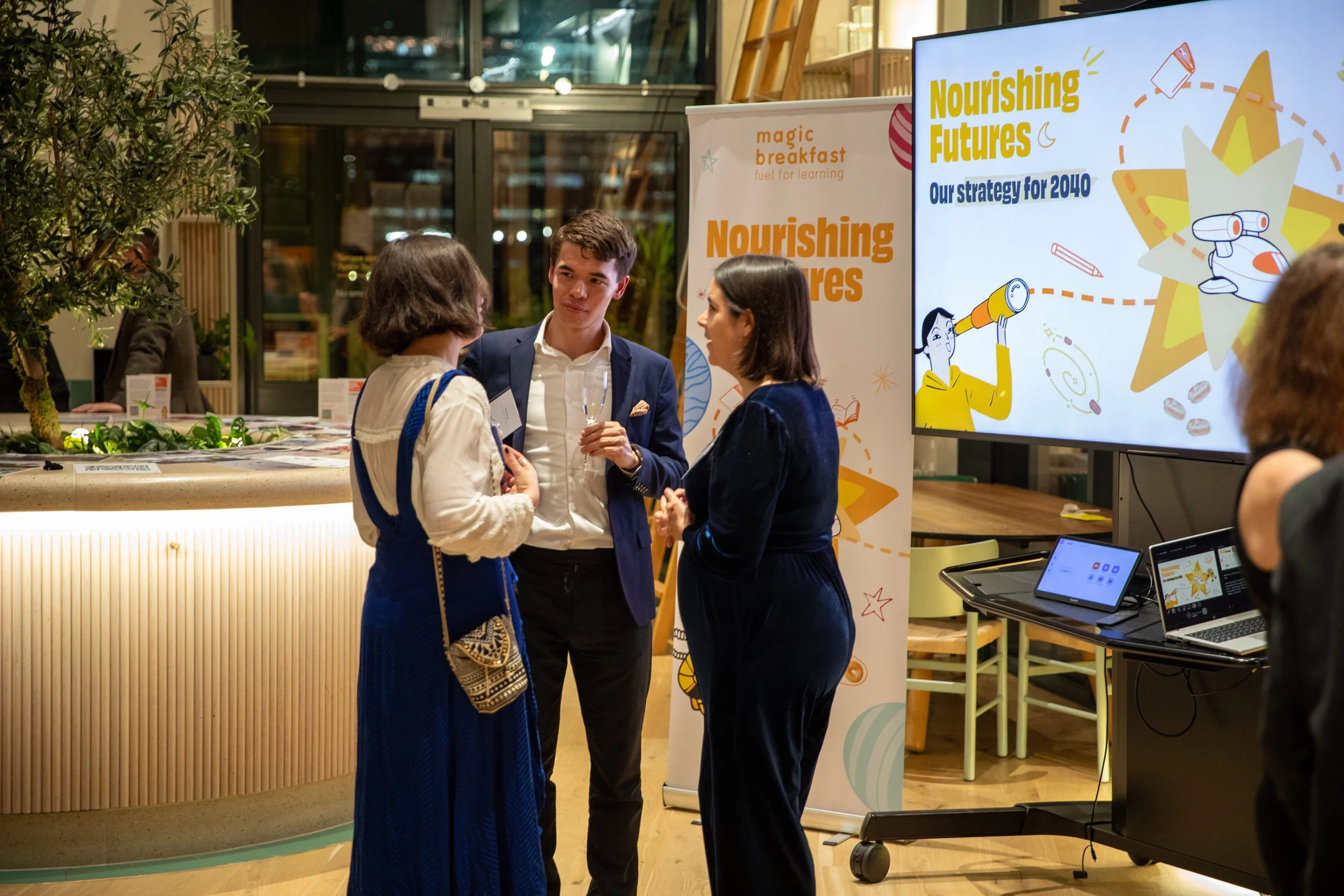 People engaged in conversation at an event, with a large digital screen displaying colorful graphics and text about future strategies and nurturing futures, in a modern, well-lit indoor setting.
