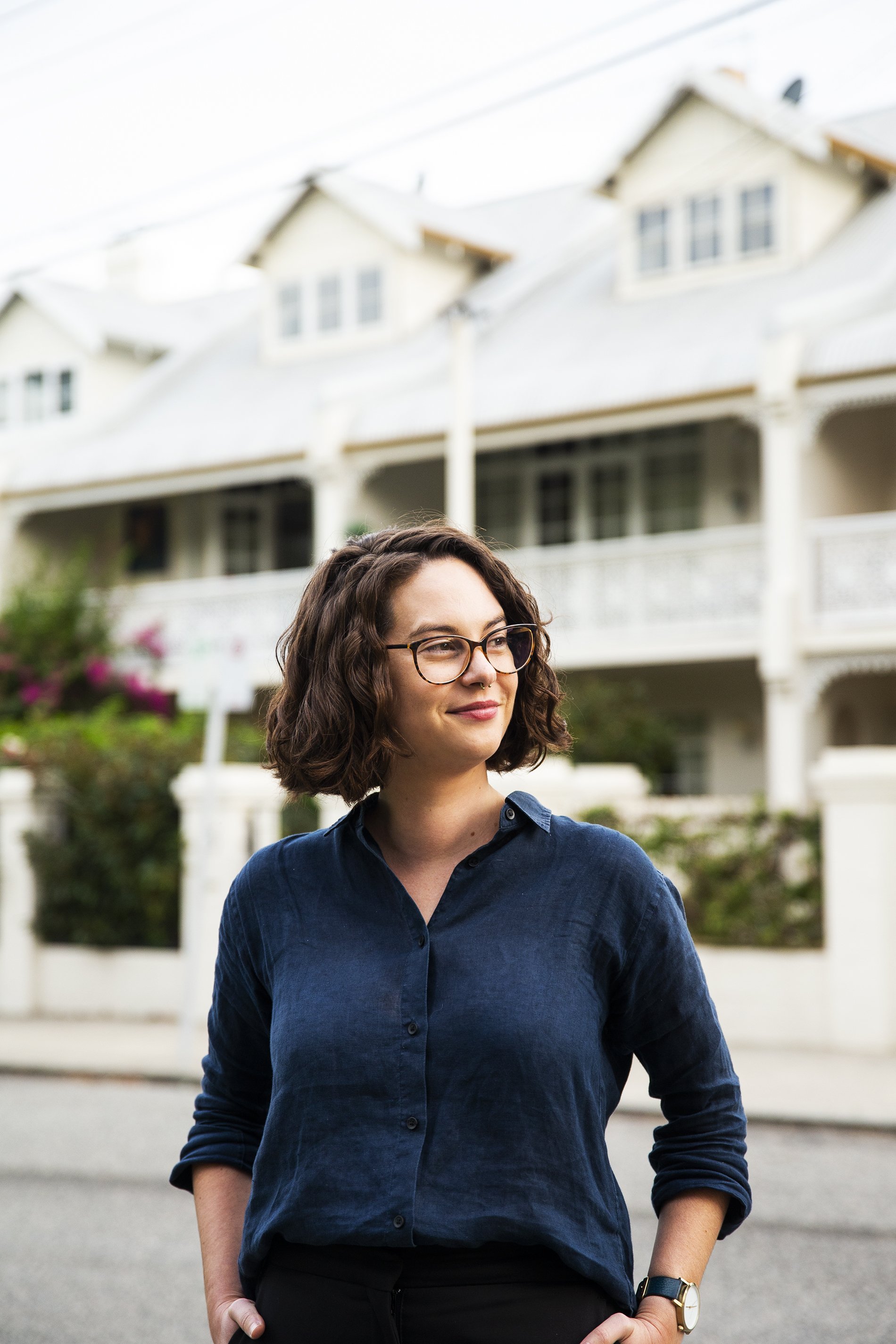 A woman with curly brown hair and glasses stands outdoors in front of a white house with multiple stories and a porch, wearing a navy blue button-up shirt and a gold wristwatch, smiling slightly.