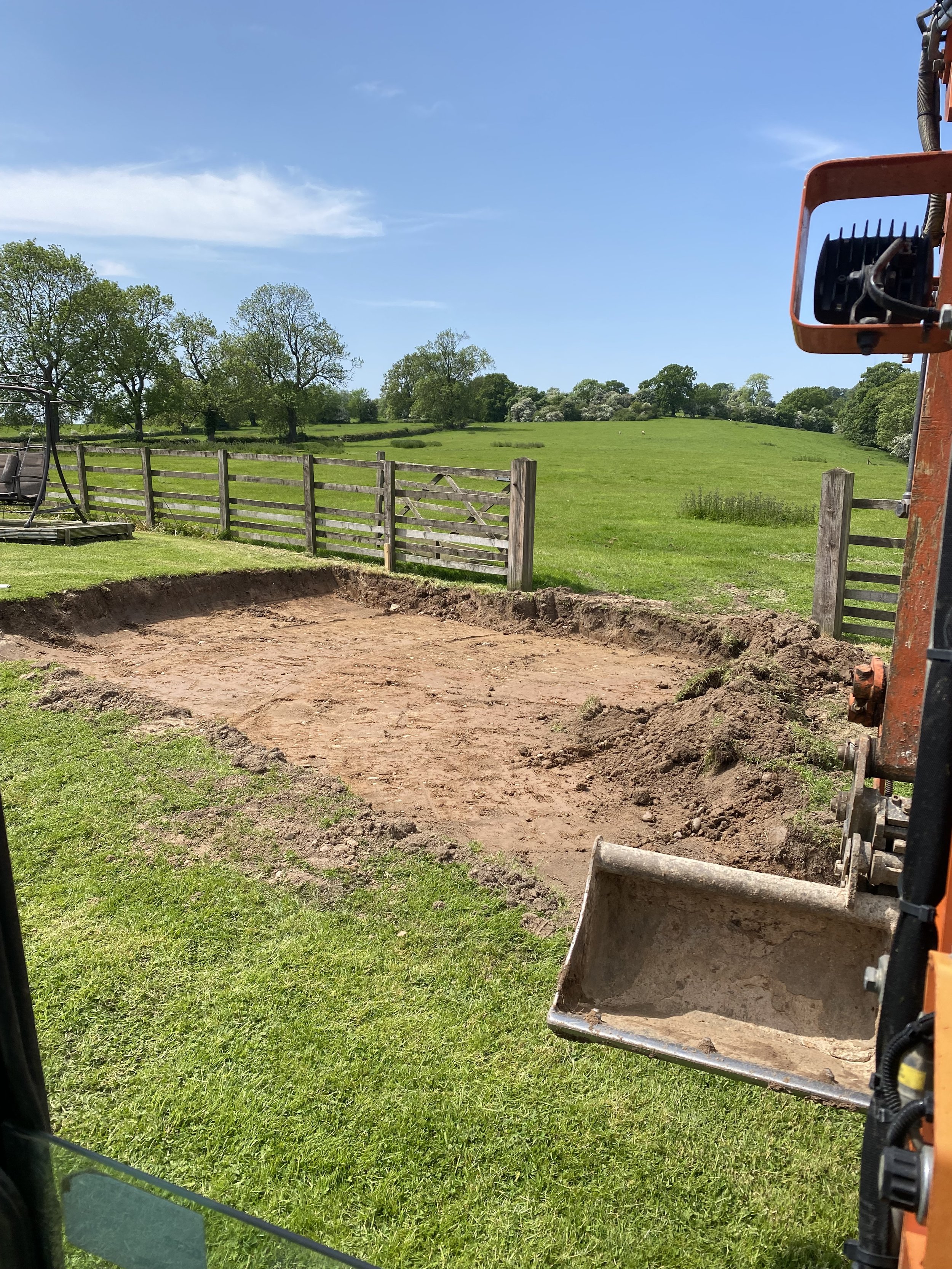 Construction site with an excavator bucket and a dug-out area on a grassy field, wooden fence, trees, blue sky with some clouds in the background.