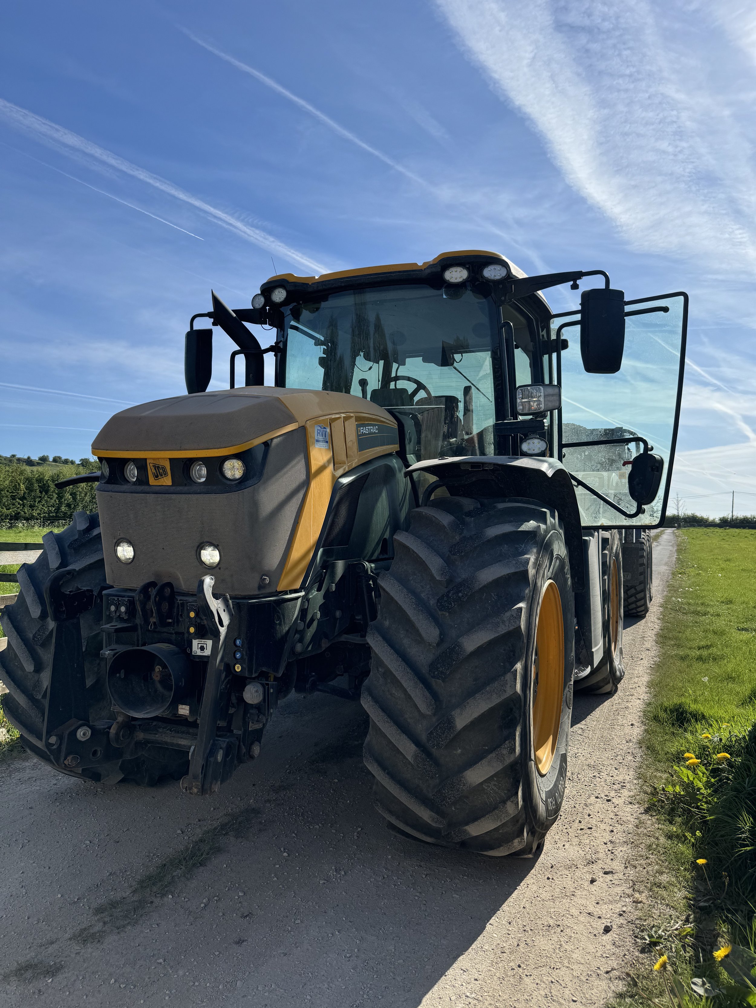 A yellow and black tractor parked on a dirt and gravel path with green grass and yellow flowers on the side under a partly cloudy sky with contrails.