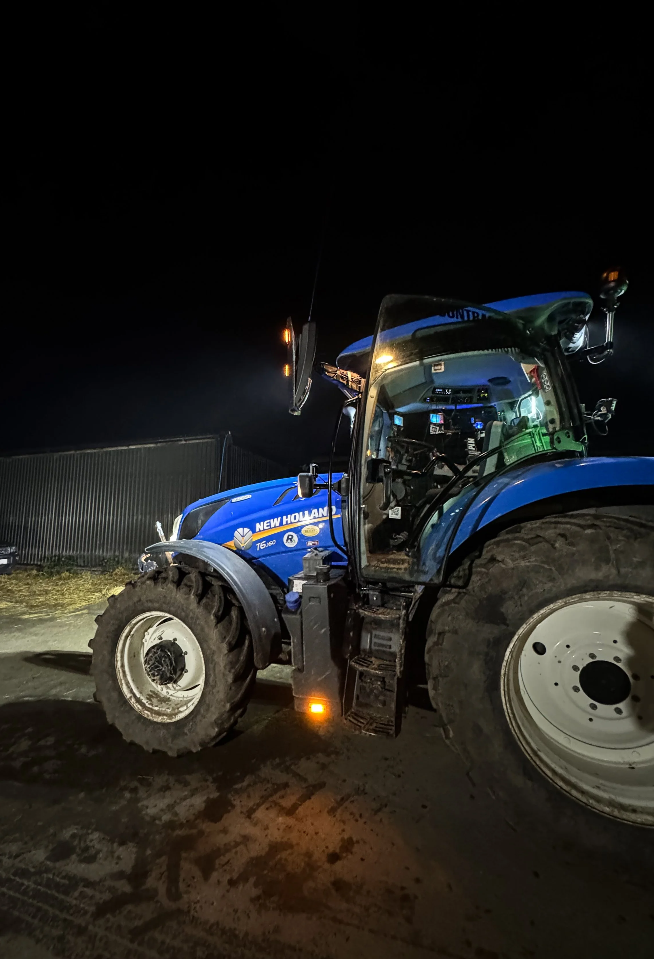 Nighttime photo of a New Holland tractor on a trailer with mud on the tires, illuminated by surrounding lights, with a fence and building in the background.