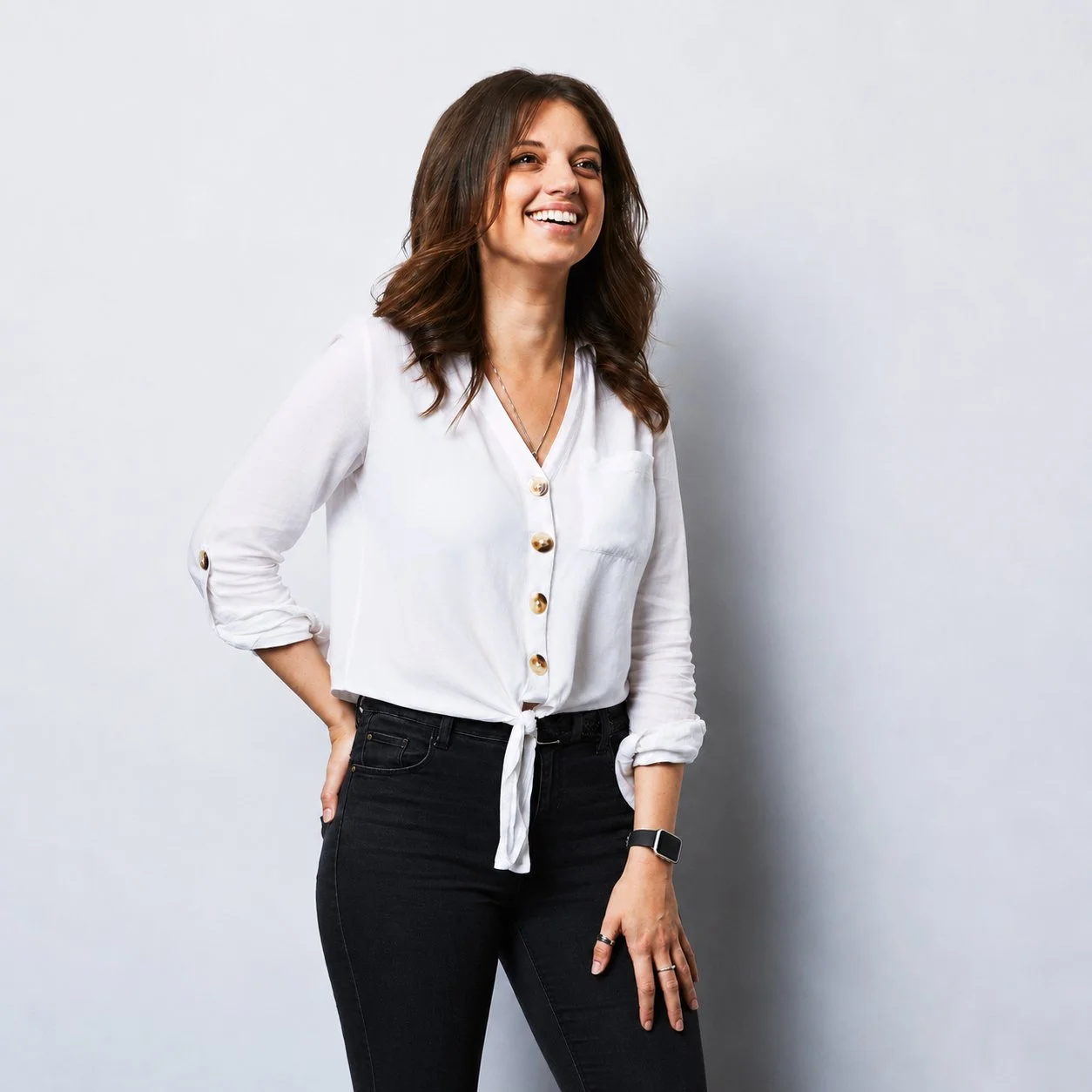 A woman with brown hair wearing a white blouse and black pants, smiling against a plain light gray background.