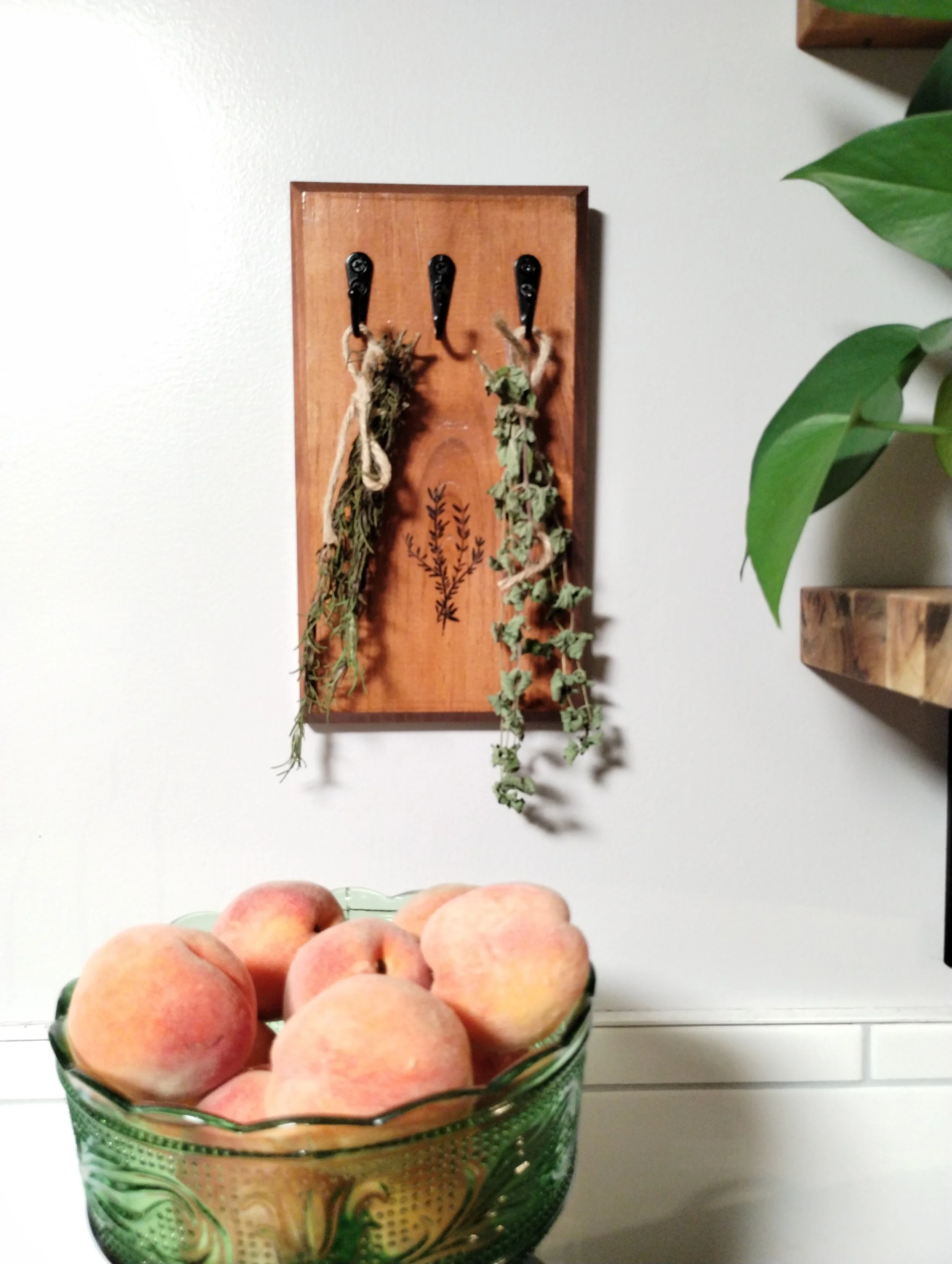Wall-mounted wooden herb drying rack with dried herbs displayed in a kitchen beside a bowl of fresh peaches