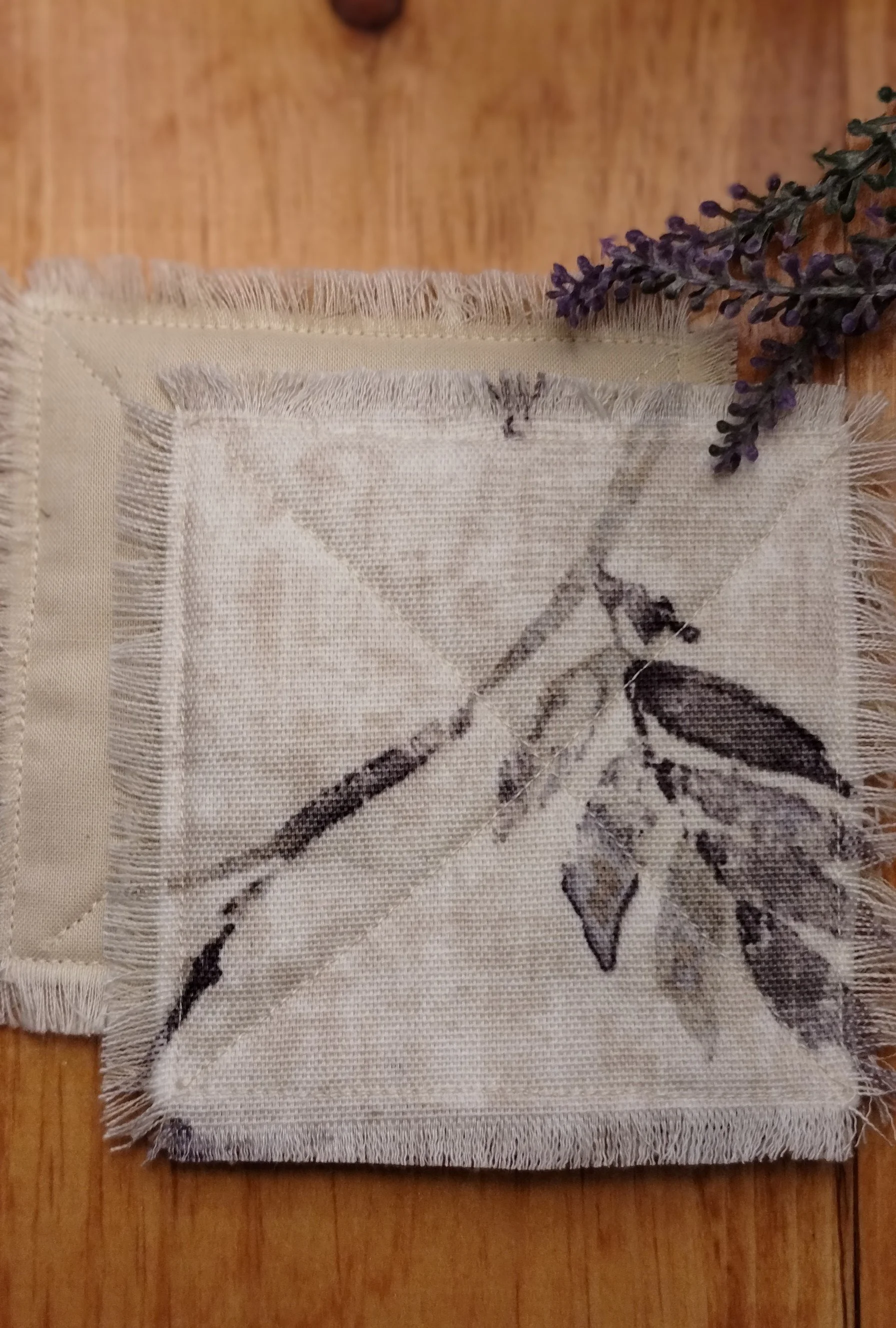 Close-up of handmade cream fabric coasters with a soft gray botanical branch print, X-stitch quilting, and frayed edges, displayed with dried lavender.