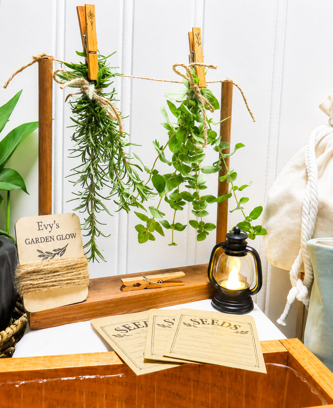 Handcrafted wooden table top herb drying rack, wood tag wrapped in jute twine, herbs hanging from drying rack attached by repurposed clothespins, 3 seed envelopes, and a mini lanter