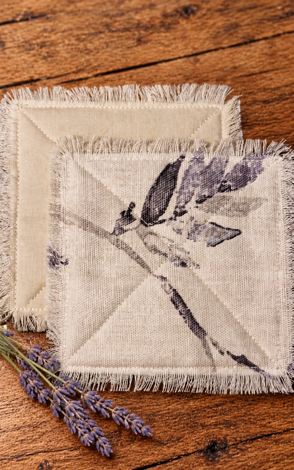 Handmade quilted fabric coasters with a muted purple and gray botanical print, natural cotton backing, and fringed edges, displayed with lavender on a wooden table.