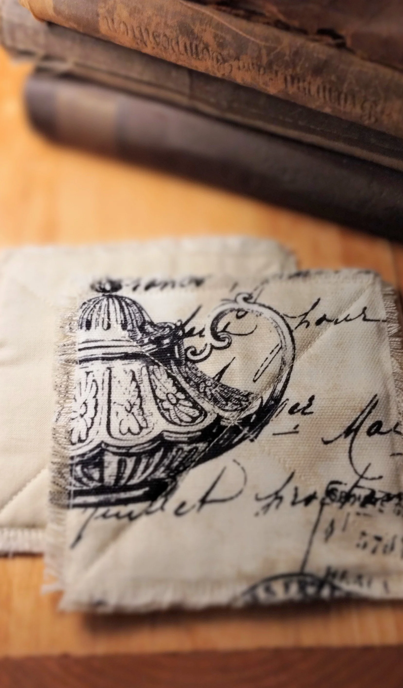 Close-up of handmade cream and black vintage script fabric coasters featuring a detailed teapot illustration, decorative stitching, and frayed edges.