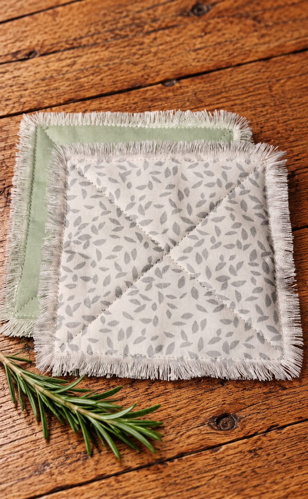 Handmade quilted fabric coasters with a delicate sage green leaf pattern, sage green backing, and fringed edges, displayed on rustic wood with fresh rosemary.