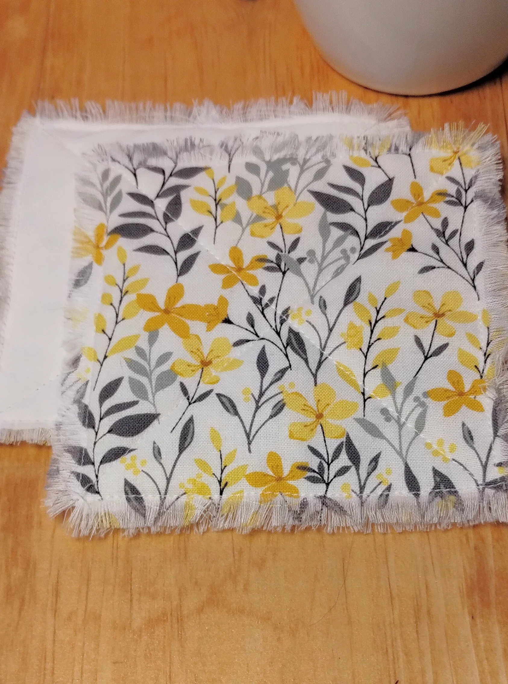 Handmade cream fabric coasters with yellow flowers and gray botanical leaves, featuring decorative X-stitch quilting and frayed edges.