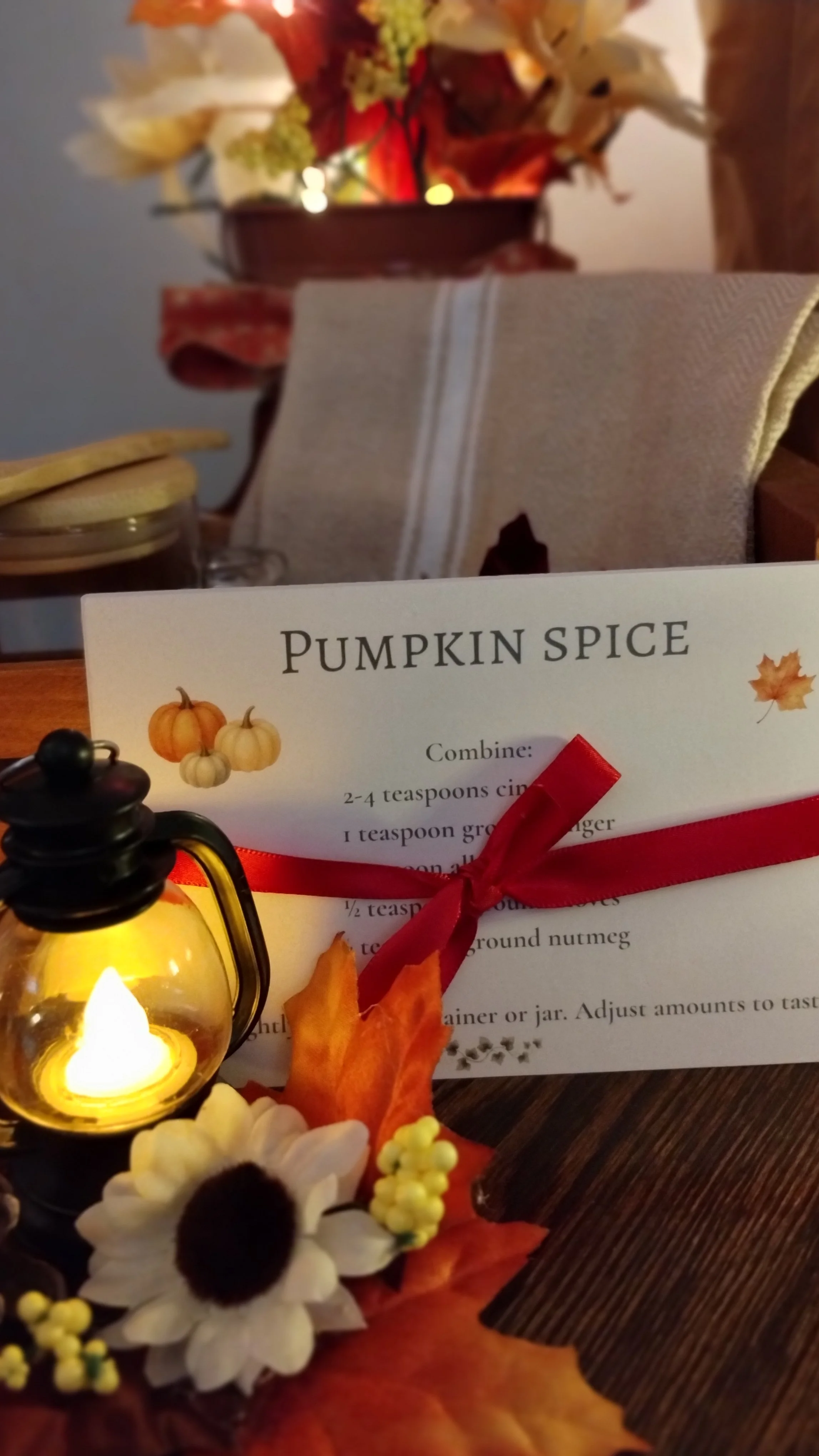 Pumpkin Spice recipe card tied with red ribbon beside a glowing mini lantern, fall leaves and cream kitchen towel.