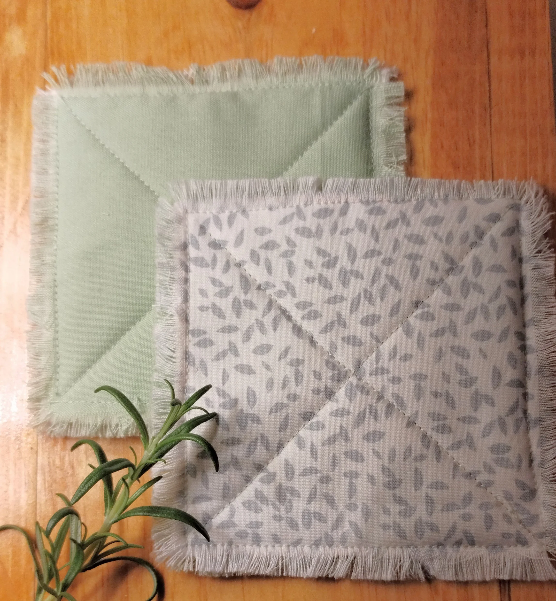 Handmade sage green and white botanical fabric coasters with decorative X-stitch quilting and frayed edges, displayed with a sprig of rosemary on a wooden table.