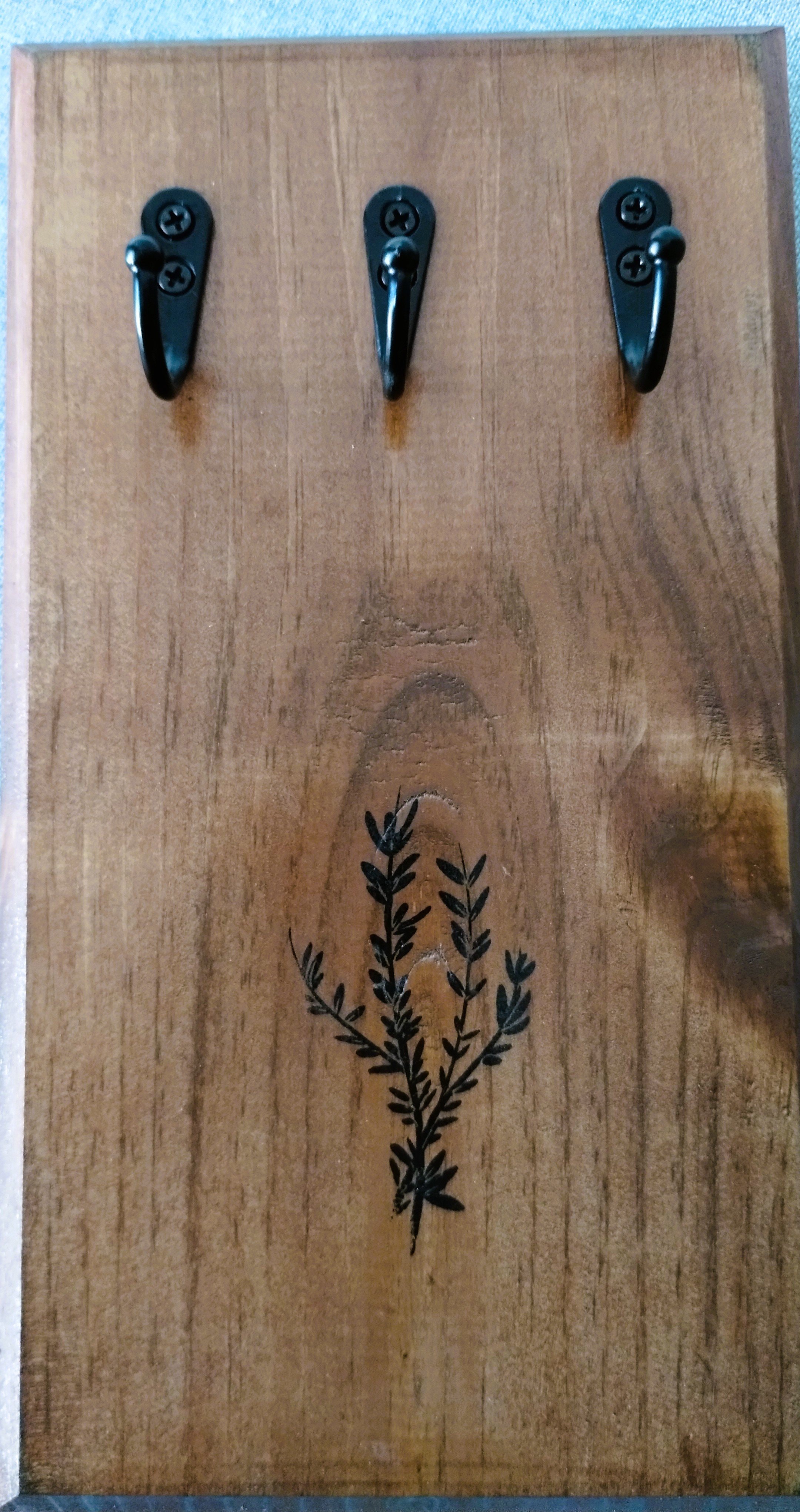 Handmade wall-mounted herb drying rack with three black hooks, warm stained pine and botanical detail.