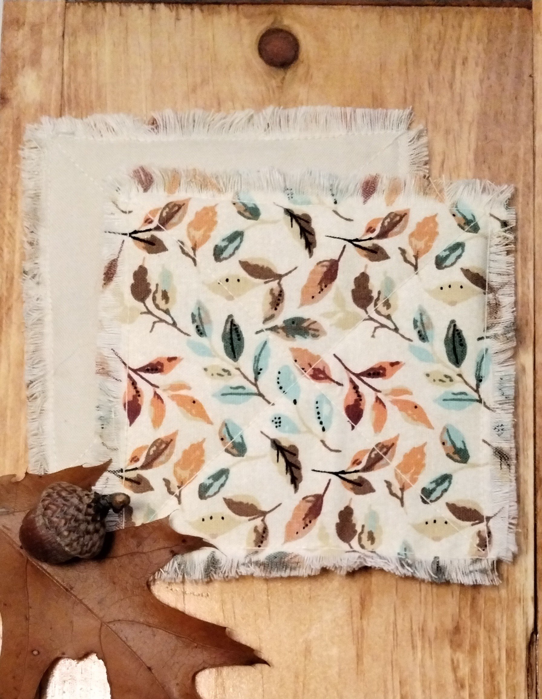 Handmade fabric coasters featuring colorful autumn leaves in shades of brown, orange, and green, with decorative X-stitch quilting and frayed edges.