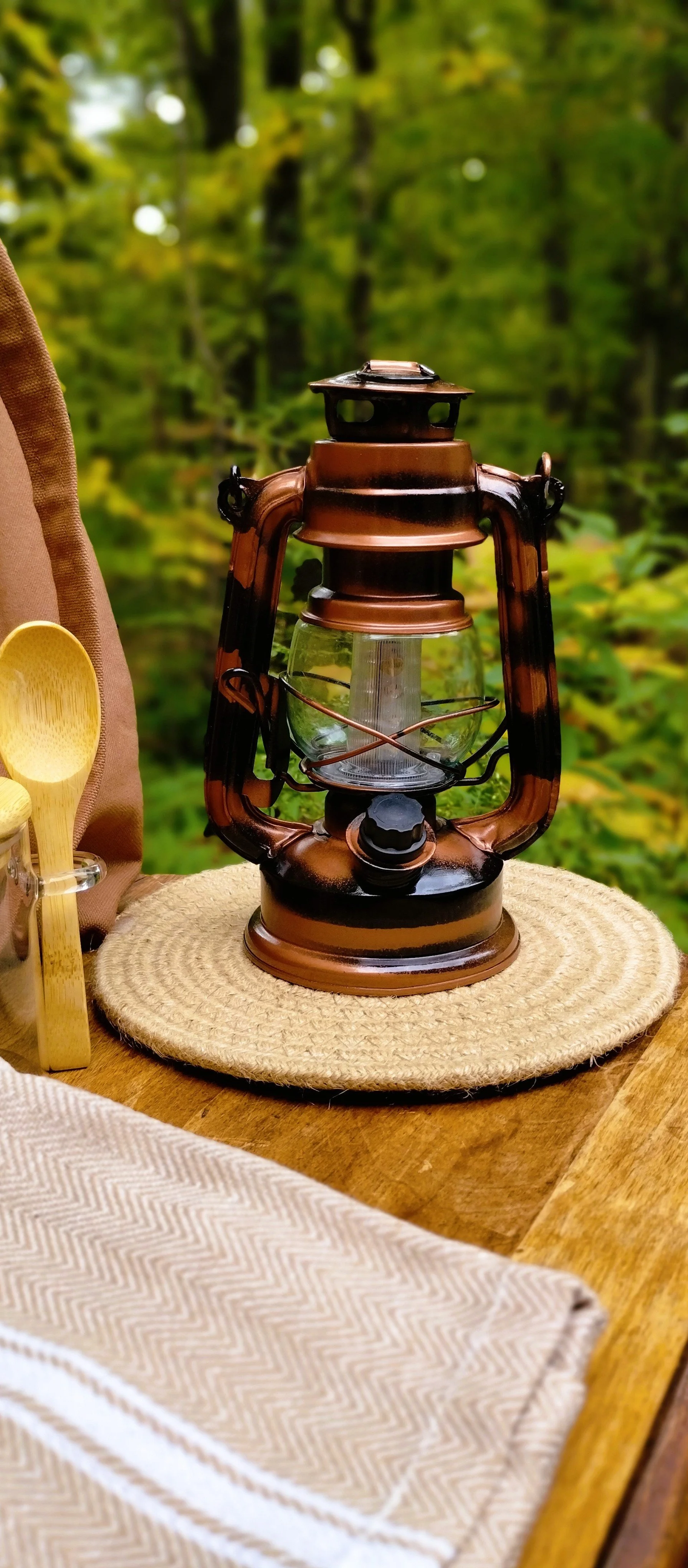 Lantern featured in Evys Garden Glow Autumn Afternoon Collection sitting on beige braided trivet with woods in the background