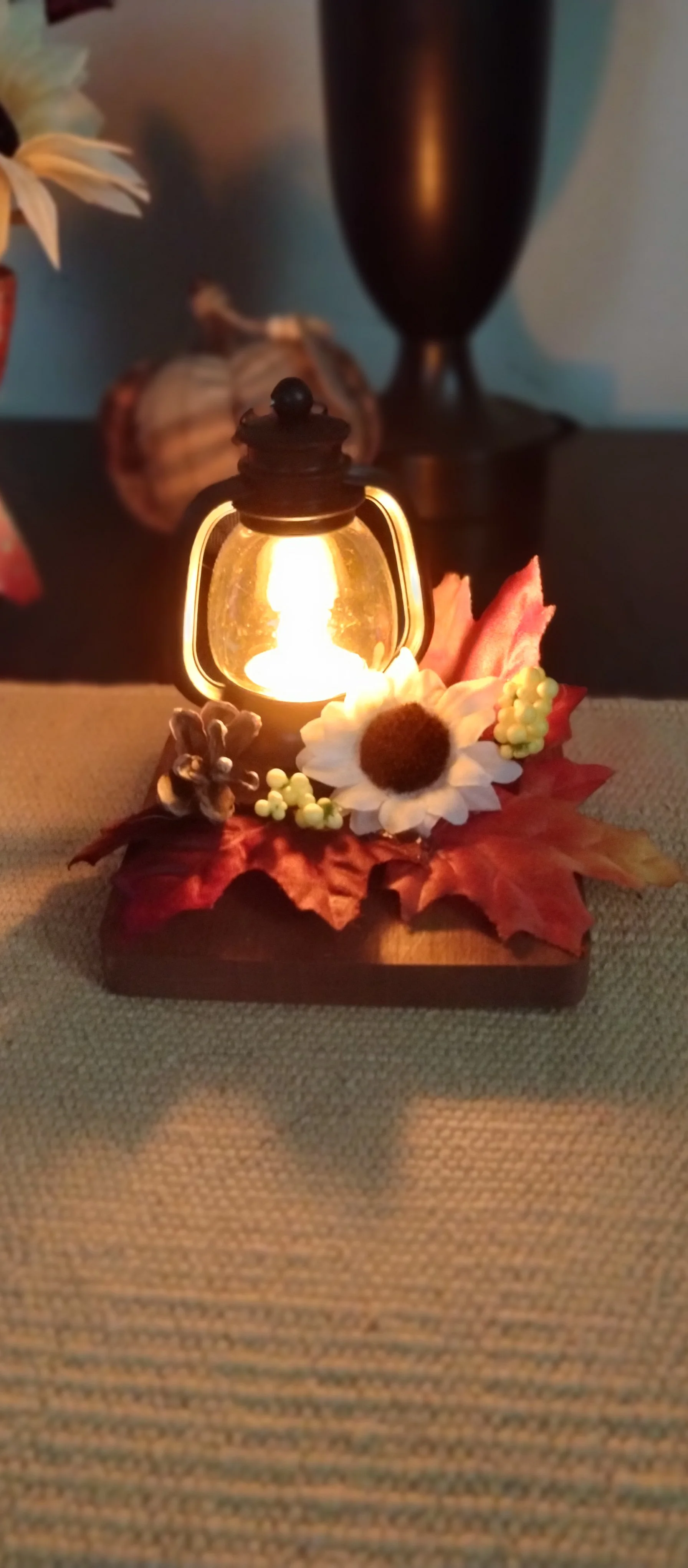 Mini lantern arrangement: Glowing mini lantern on a stained wooden coaster decorated with fall leaves, white flower, berries, and pinecone.