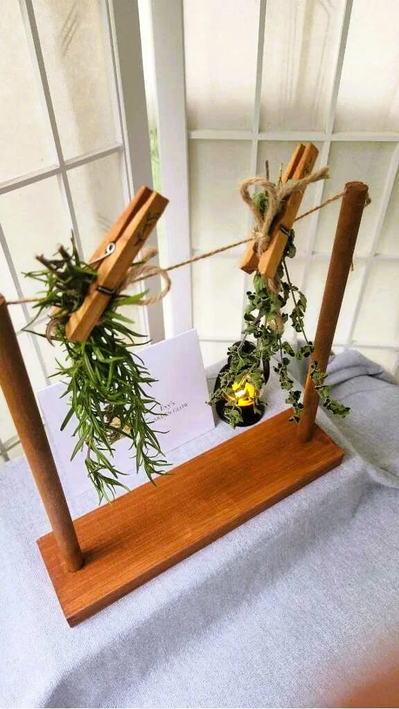 Fresh rosemary and herbs hanging from wooden clothespins on a tabletop wooden herb drying rack near a window.