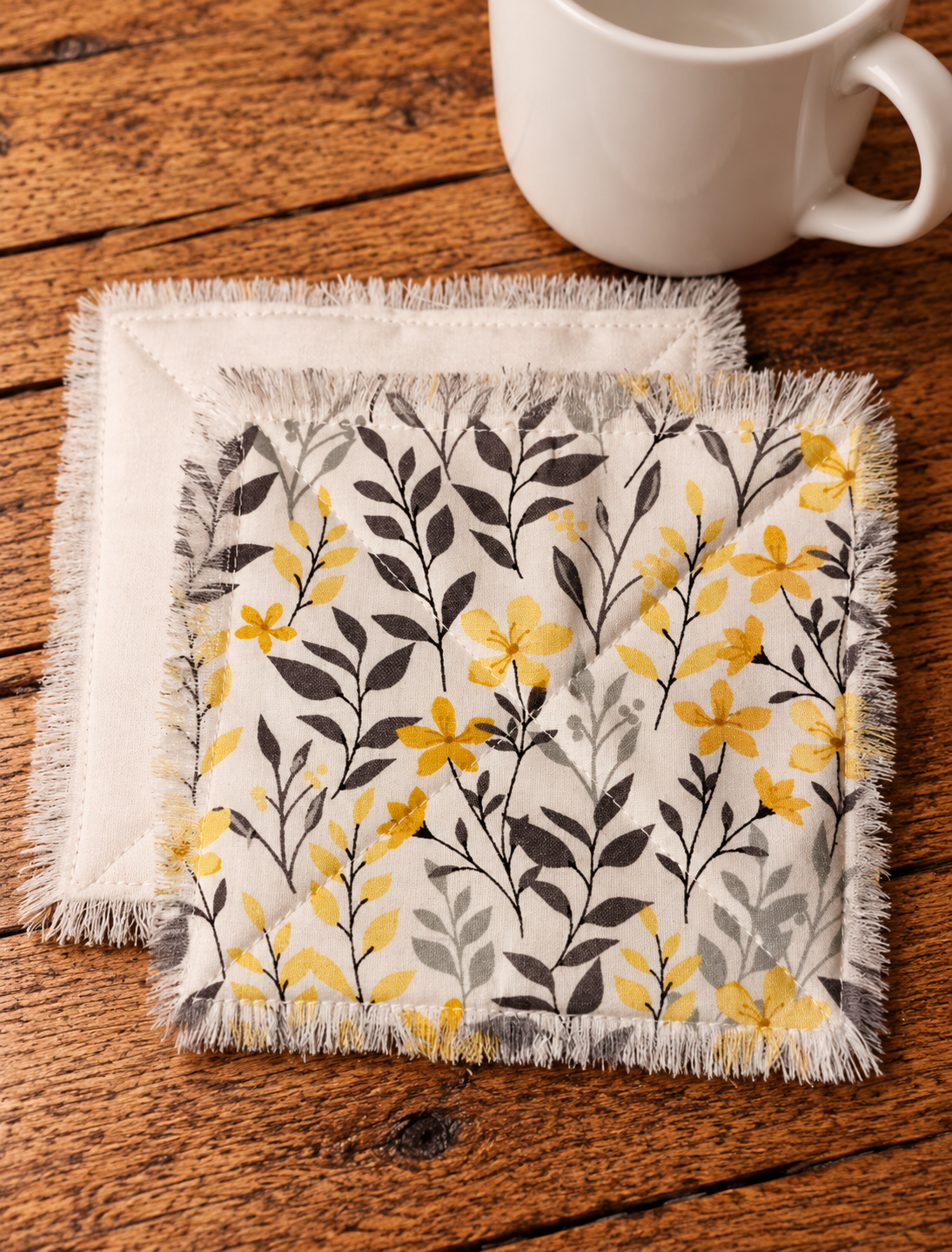 Handmade fabric coasters featuring yellow flowers and charcoal gray leaves on cream fabric, with quilted stitching and fringed edges beside a white ceramic mug.