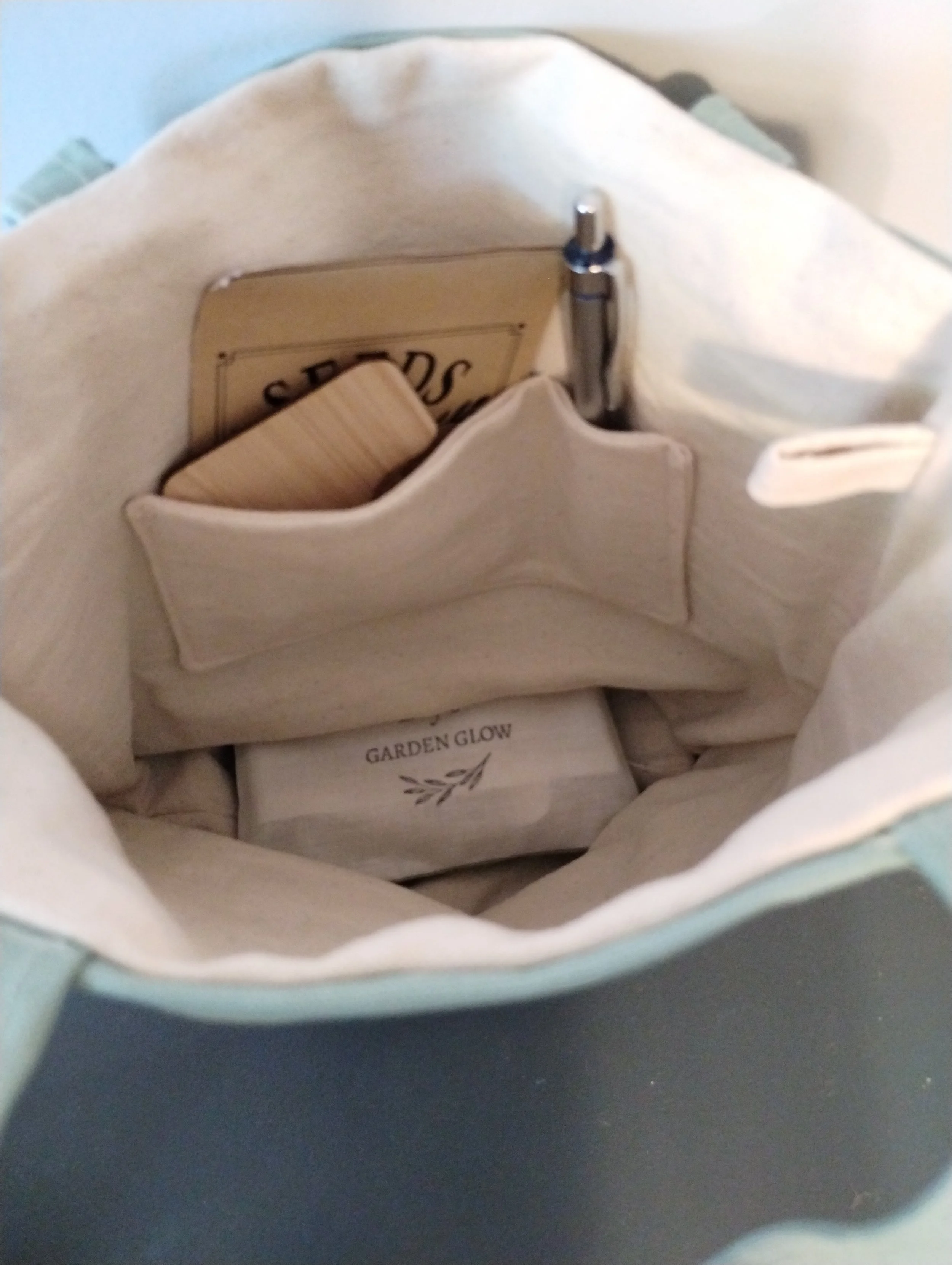 Inside view of the sage green tote, featuring 2 pockets showing one holding a pen and the other with seed envelopes and plant markers, and a key loop