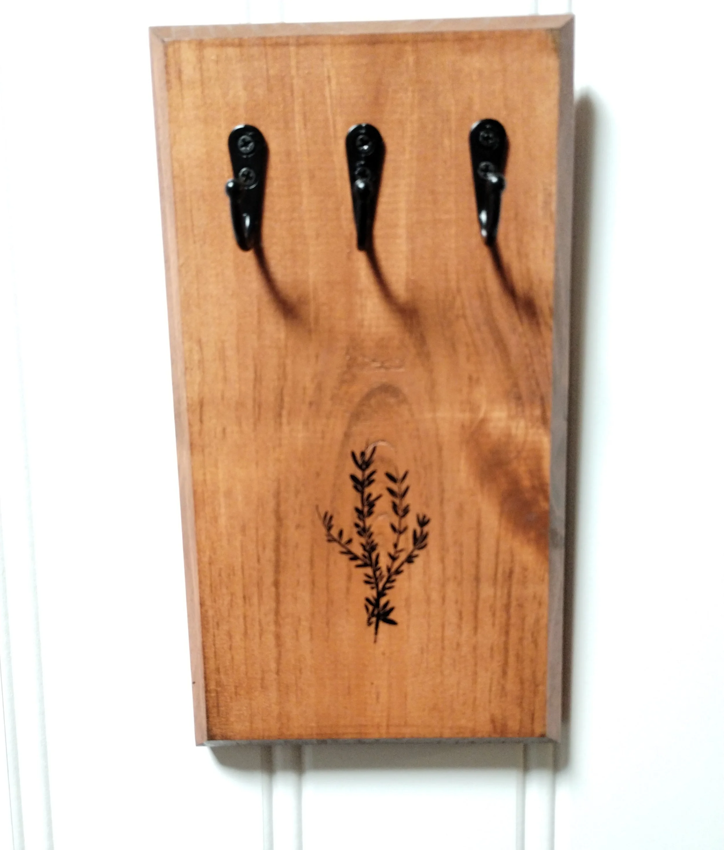 Front view of wooden wall-mounted herb drying rack with three matte black hooks and stamped botanical design