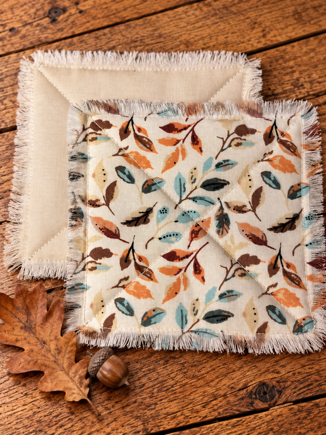 Handmade quilted fabric coasters with rust, teal, and brown leaf patterns, fringed edges, and natural cotton backing on a rustic wooden table with an oak leaf and acorn.