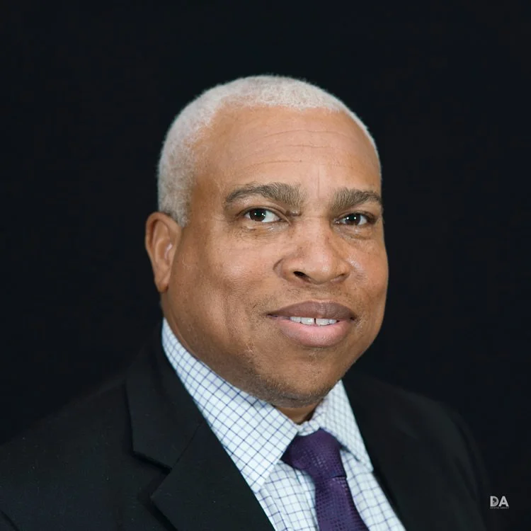 Portrait of a middle-aged African American man with short gray hair, wearing a dark suit, a checkered dress shirt, and a purple tie against a black background.