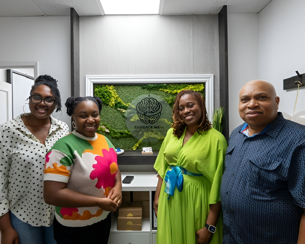 Four people standing in an office, smiling, in front of a green company logo and sign on the wall.