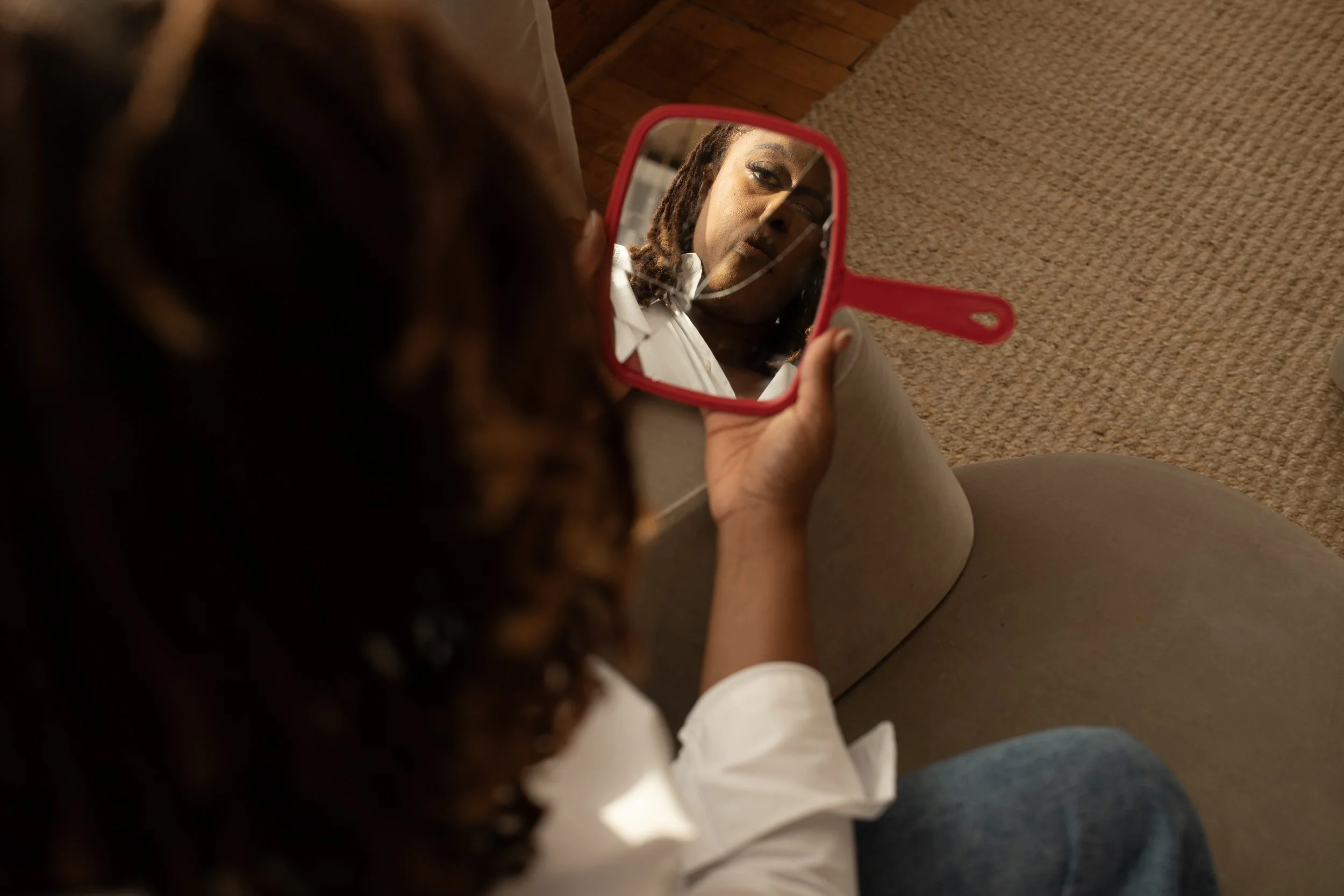 Person holding a small hand mirror, looking at their reflection on a brown carpeted floor.