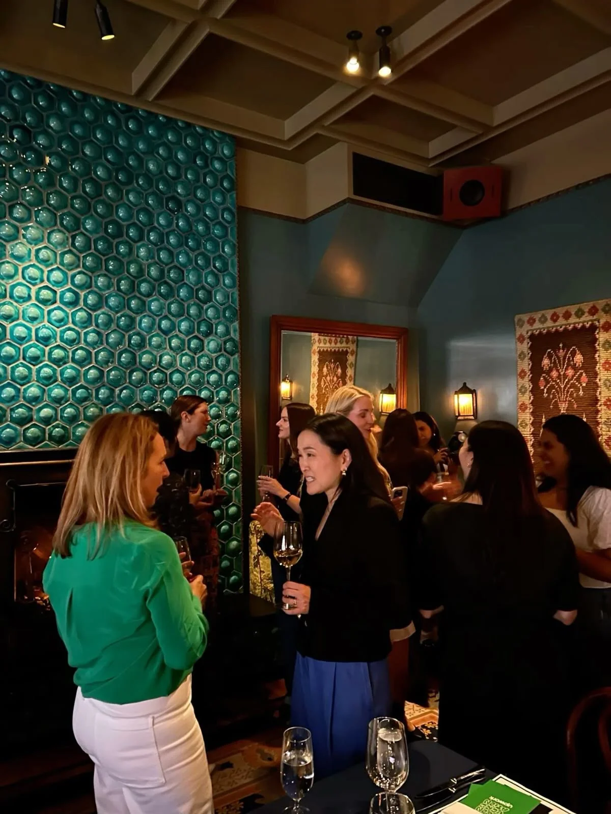 Group of women socializing in a restaurant or bar with dark walls, decorative tiles, and warm lighting.