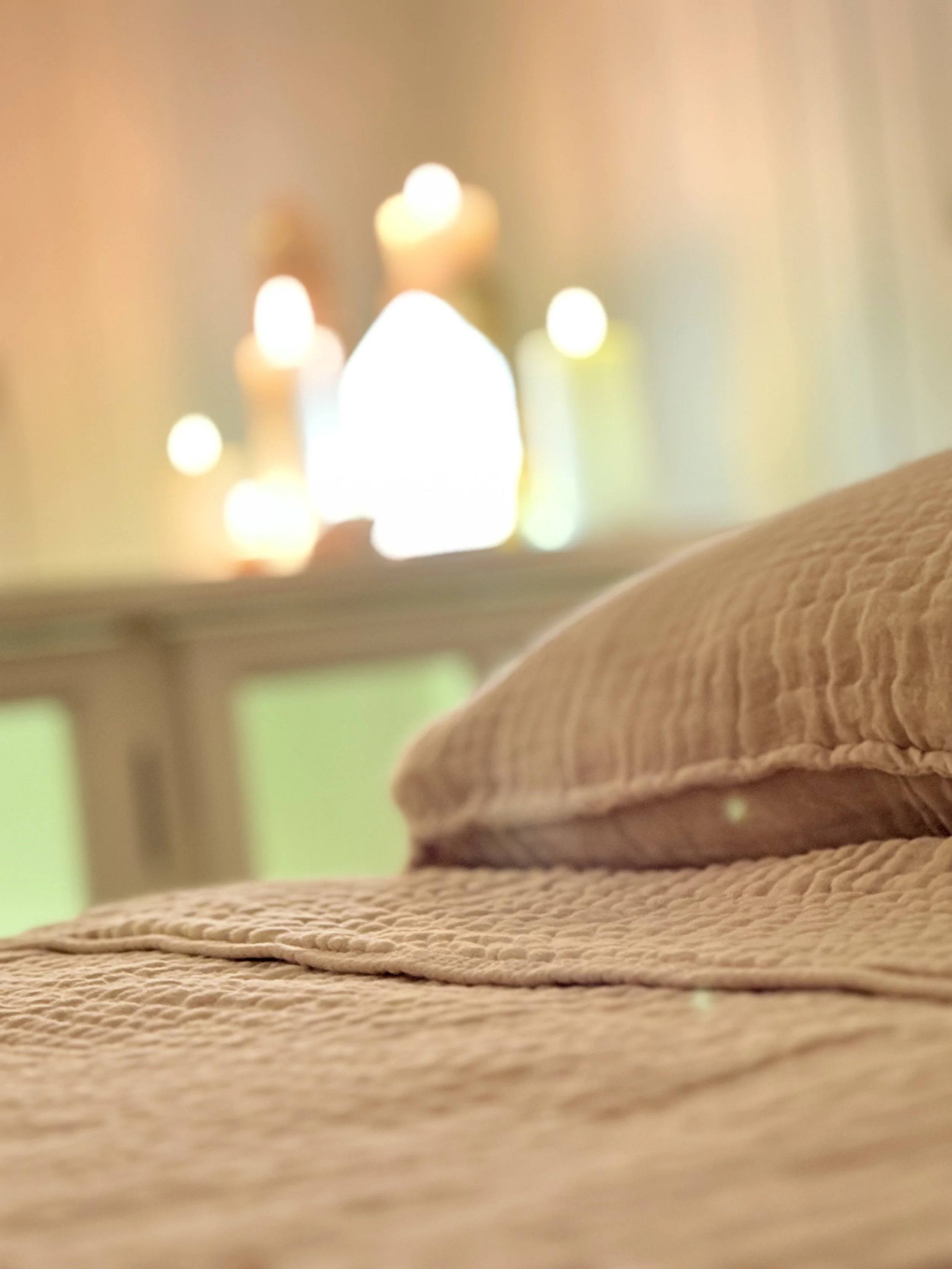 Close-up of a beige woven fabric surface with a folded cloth on top in the foreground. Out-of-focus background features several lit candles emitting a soft, warm glow.