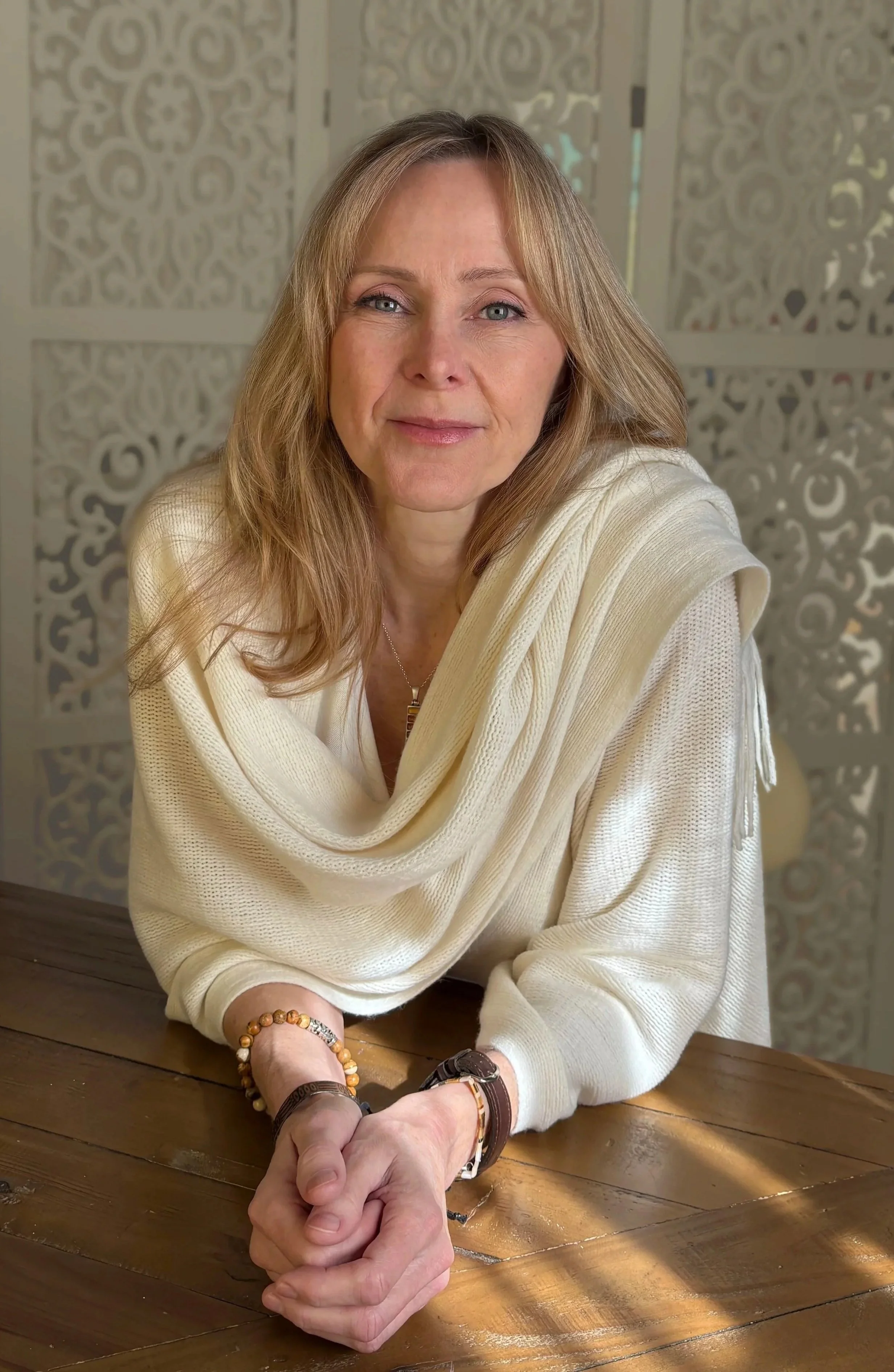 A middle-aged woman with long blonde hair, wearing a cream-colored sweater, sitting at a wooden table with her hands clasped. She is indoors with decorative white lattice room dividers in the background.
