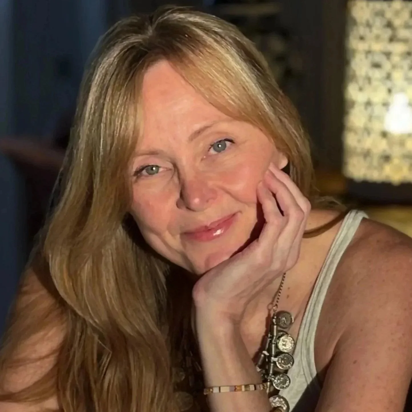 A woman with long red hair and blue eyes resting her chin on her hand, smiling softly at the camera. She is wearing a white tank top and accessories including a bracelet and necklace. The background appears to be indoors with dim lighting.