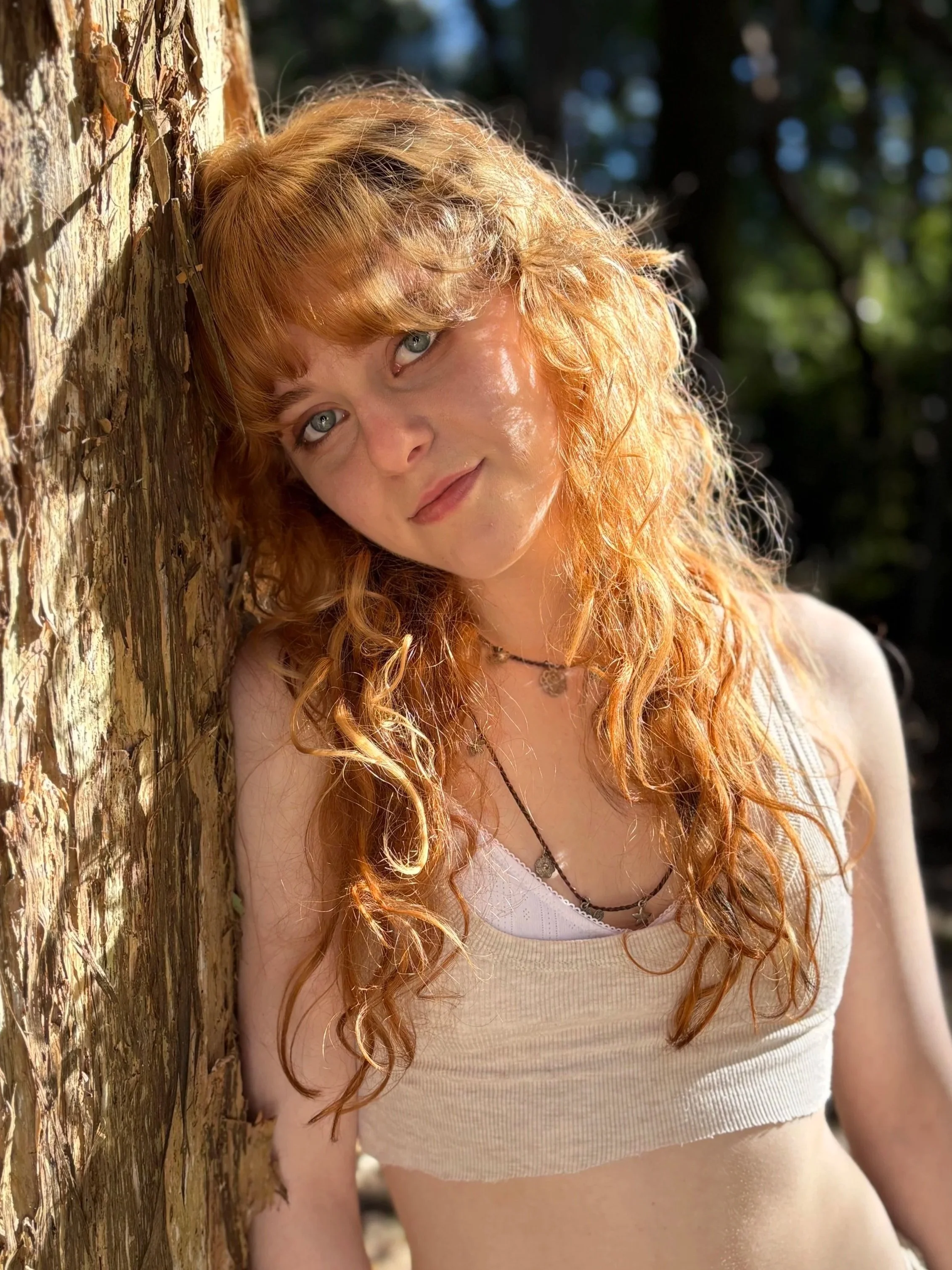 A young woman with long, curly red hair and blue eyes leaning against a tree outdoors. She is wearing a light-colored sleeveless top and a necklace, with a blurred natural background.