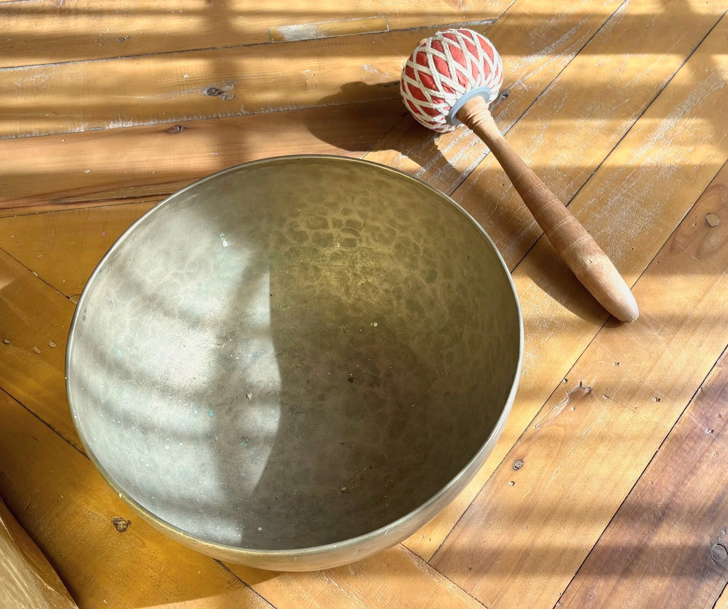 A metal handpan drum with a wooden stick resting on it, placed on a wooden floor.