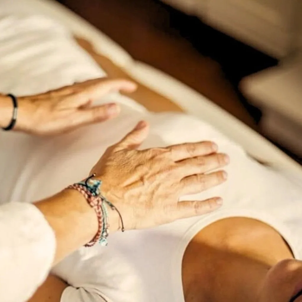 Close-up of a person's elderly hands resting on their thigh, showing multiple bracelets and a black wristband, next to a young person's arm and leg, on a bed.