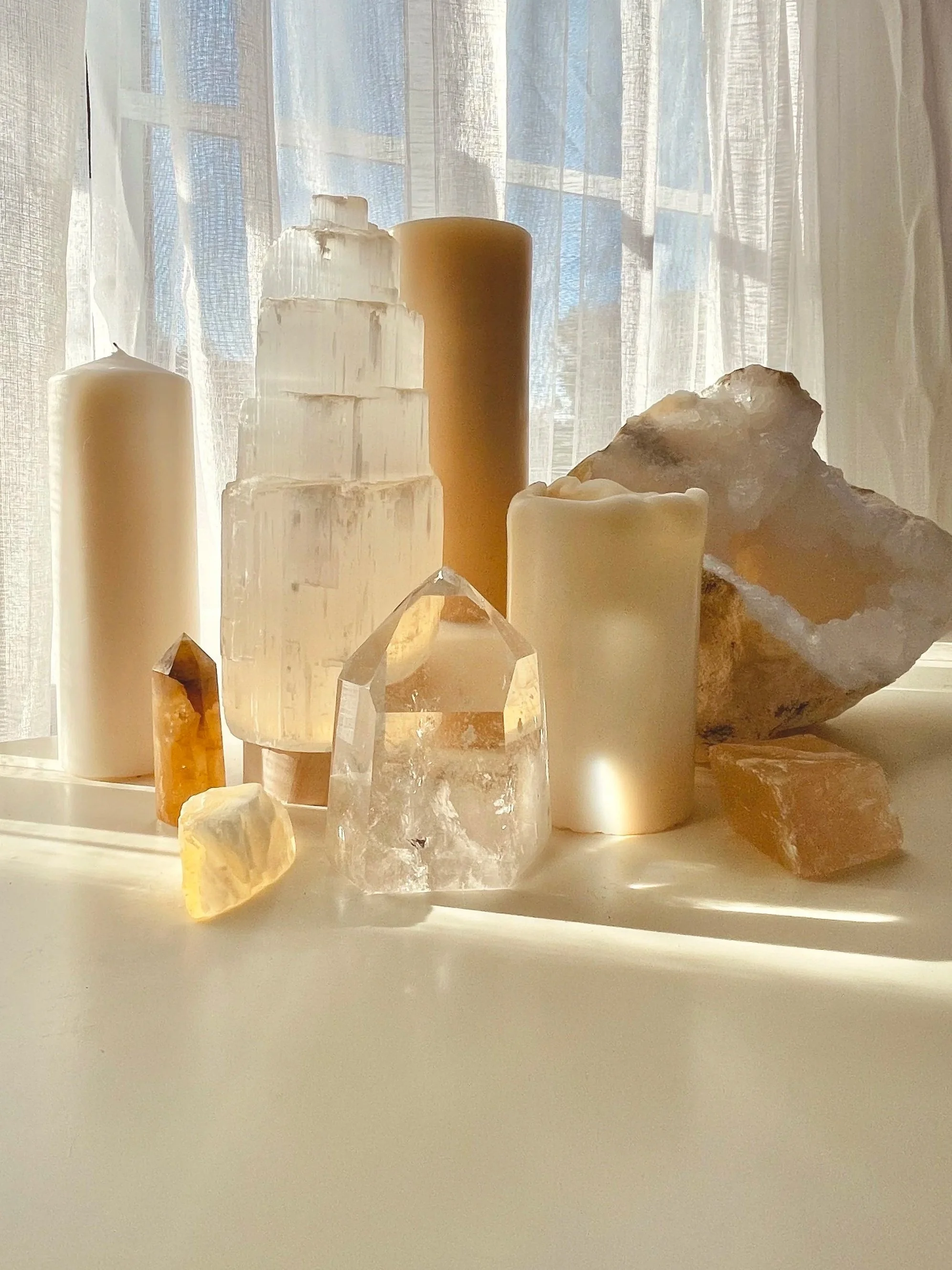 Various candles and crystals on a white surface with sheer curtains and a window in the background.