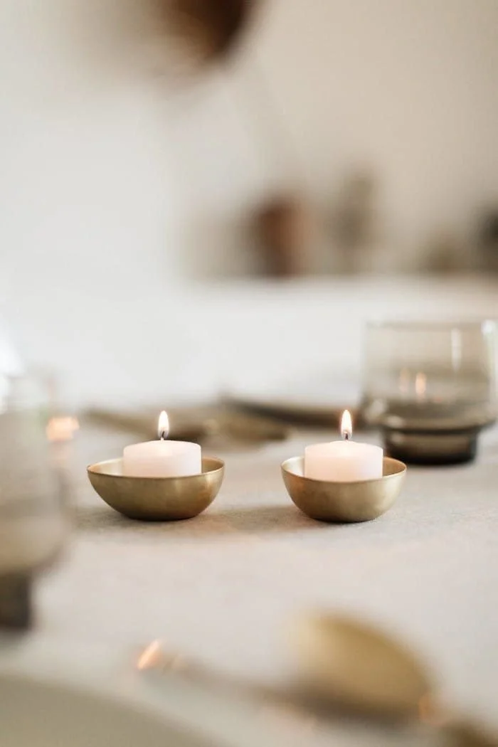 Two lit white candles in gold-colored bowls on a table with blurred background.