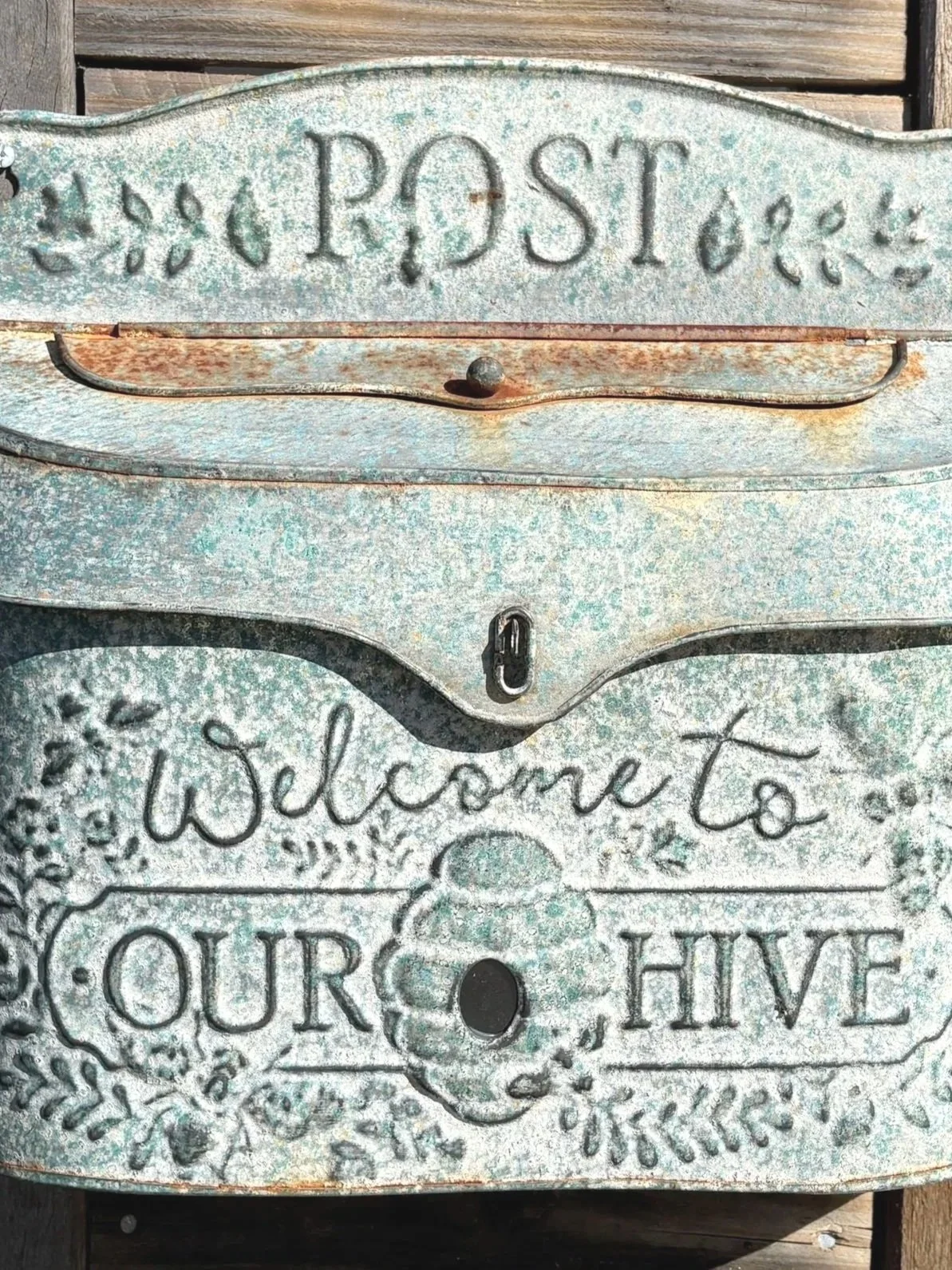 A vintage, weathered mailbox with a decorative design and the words 'Welcome to our hive.' The mailbox has a rusty lock and a slot for mail, with the top sign reading 'Post' in capital letters.