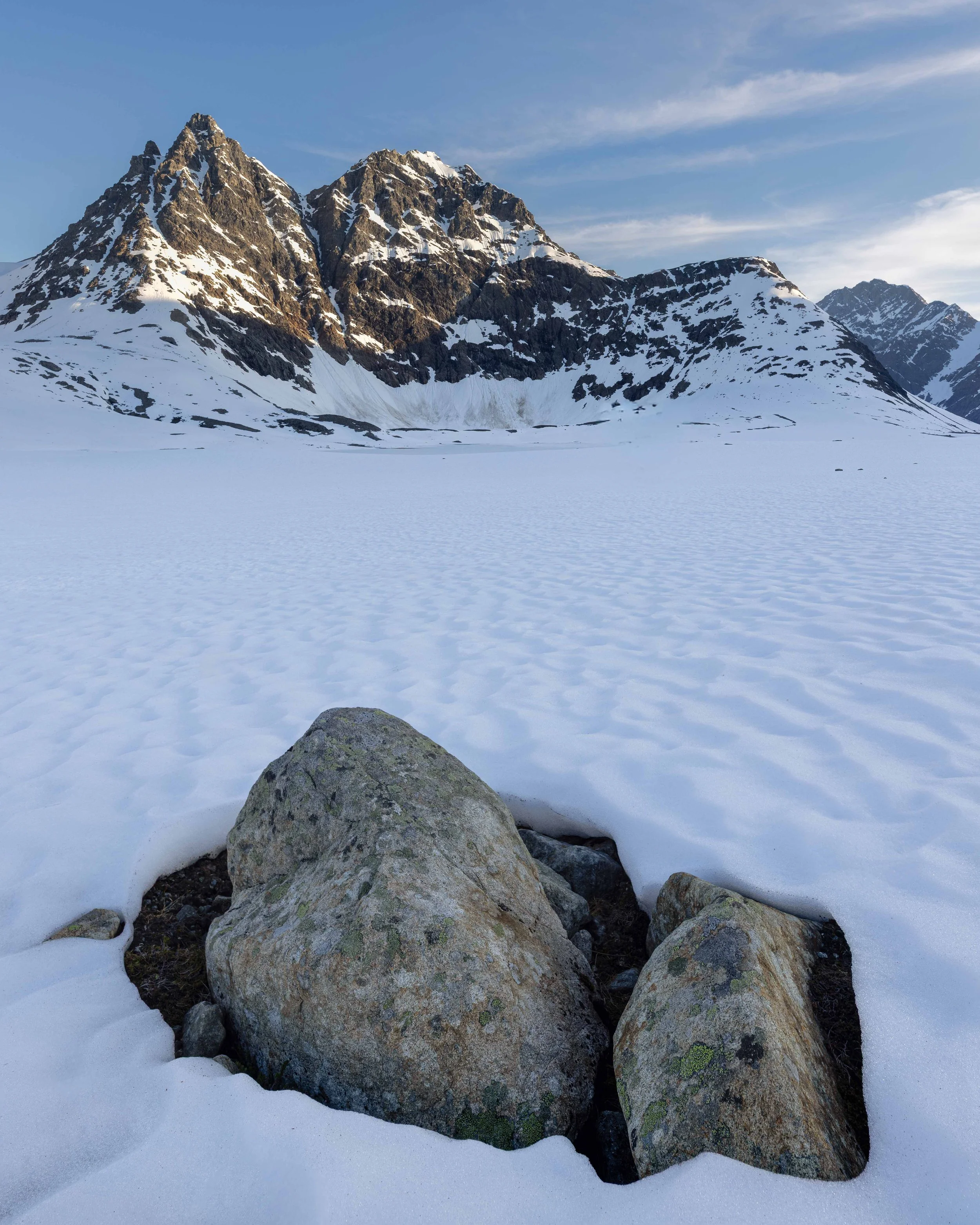 Fotografía vertical de paisaje invernal con una roca en primer plano, nieve texturizada y montañas escarpadas en el norte de Noruega.