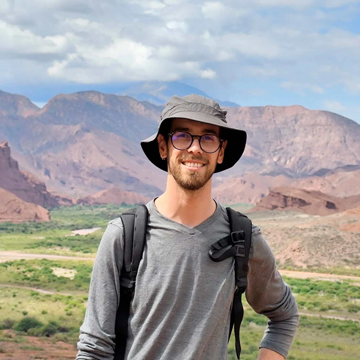 Hombre con gafas, sombrero y mochila, sonriendo, en un paisaje de montañas con cielo nublado.