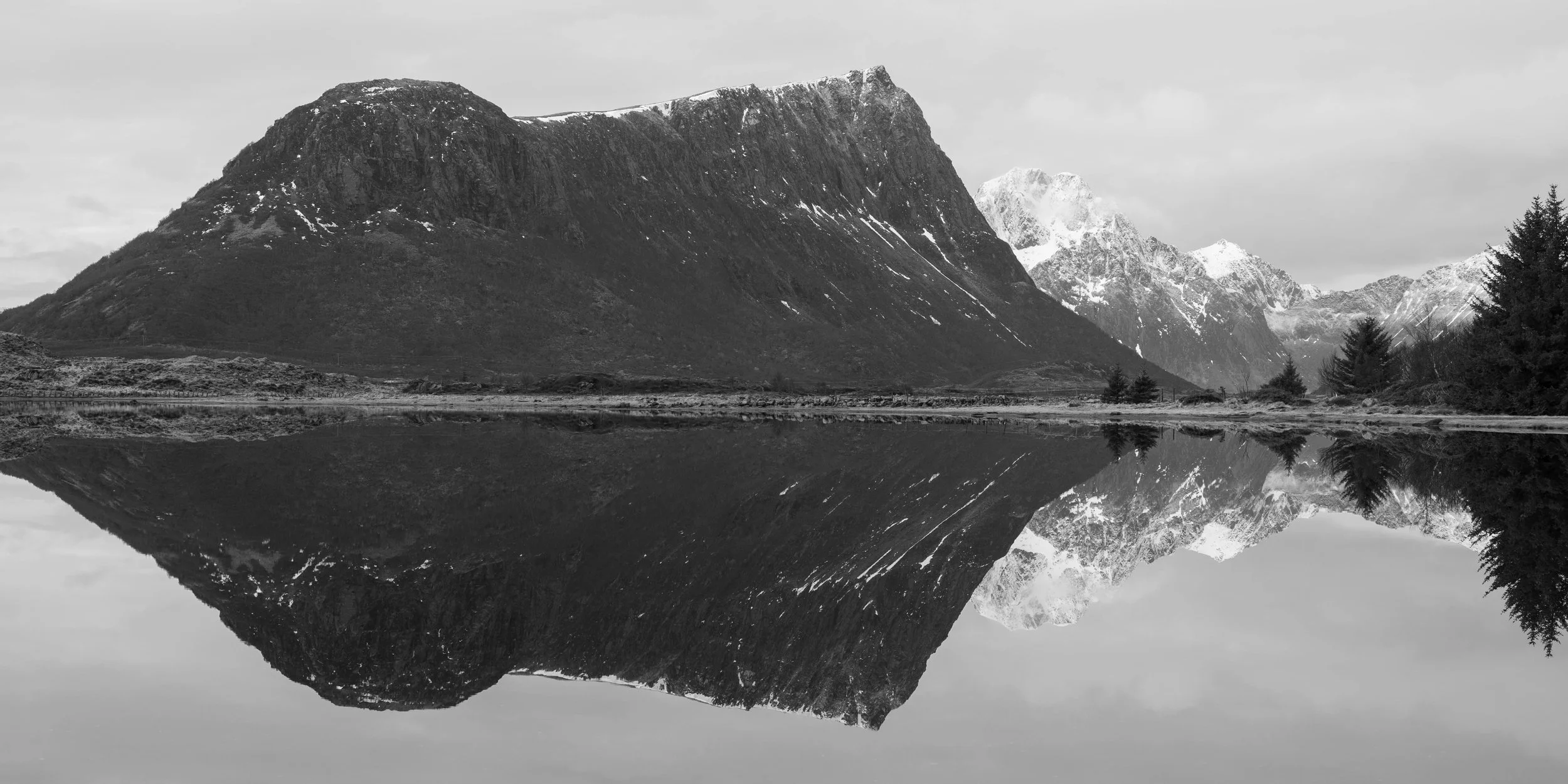 Fotografía minimalista en blanco y negro de montañas en Lofoten Noruega reflejadas sobre un lago o fiordo en calma, creando una composición simétrica y serena.