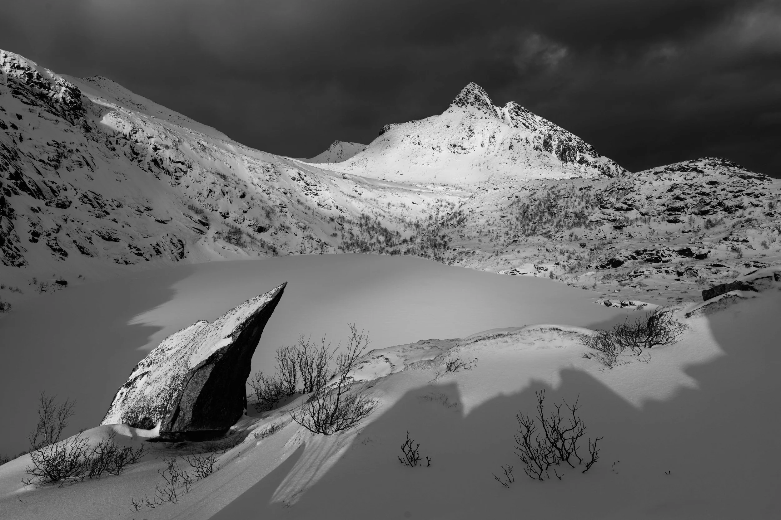 Imagen en blanco y negro de un paisaje invernal con montañas nevadas, lago congelado, rocas en primer plano y un cielo oscuro lleno de contraste. Lofoten