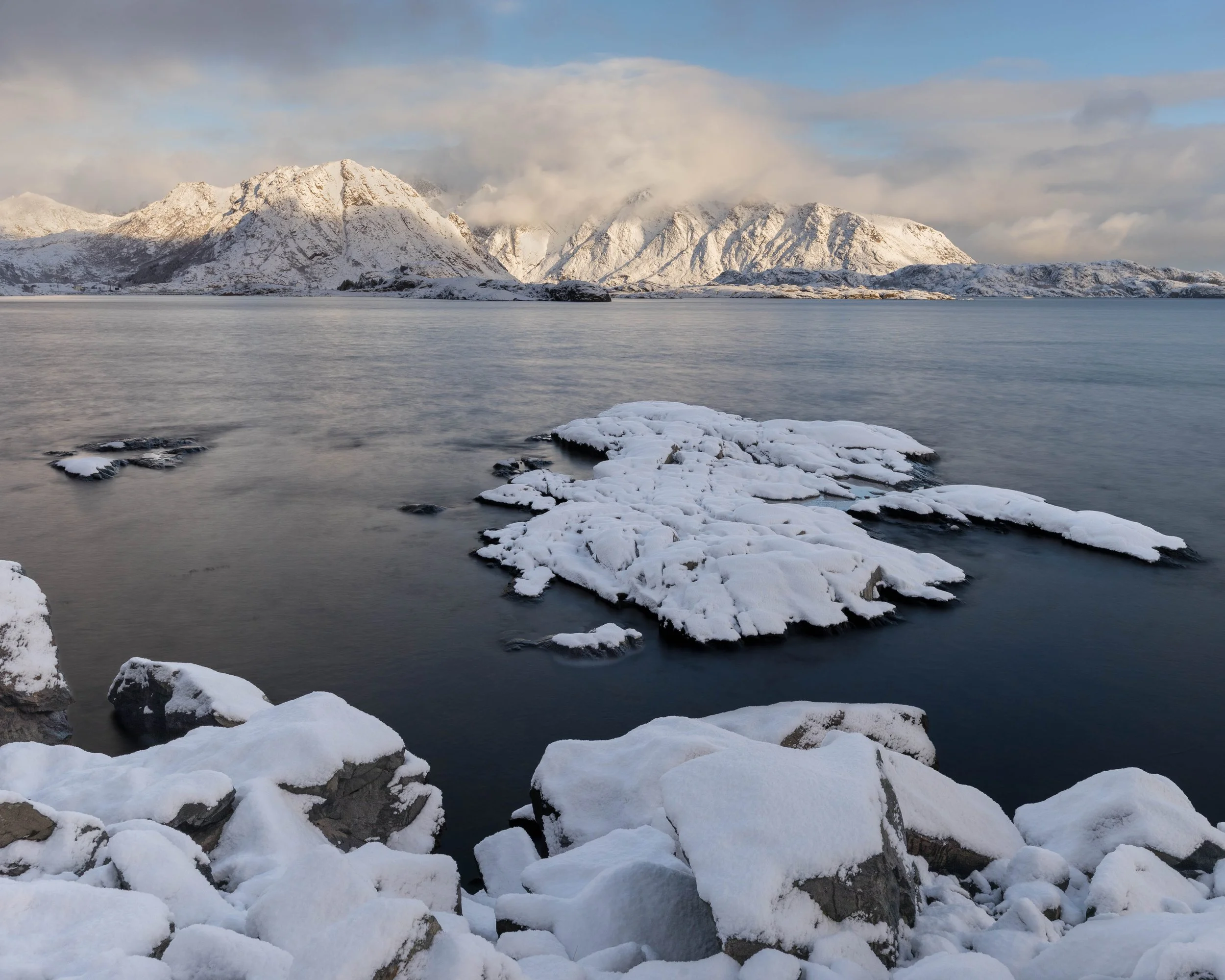 Paisaje invernal en Lofoten norte de Noruega con rocas cubiertas de nieve, aguas calmas y montañas iluminadas suavemente entre nubes.