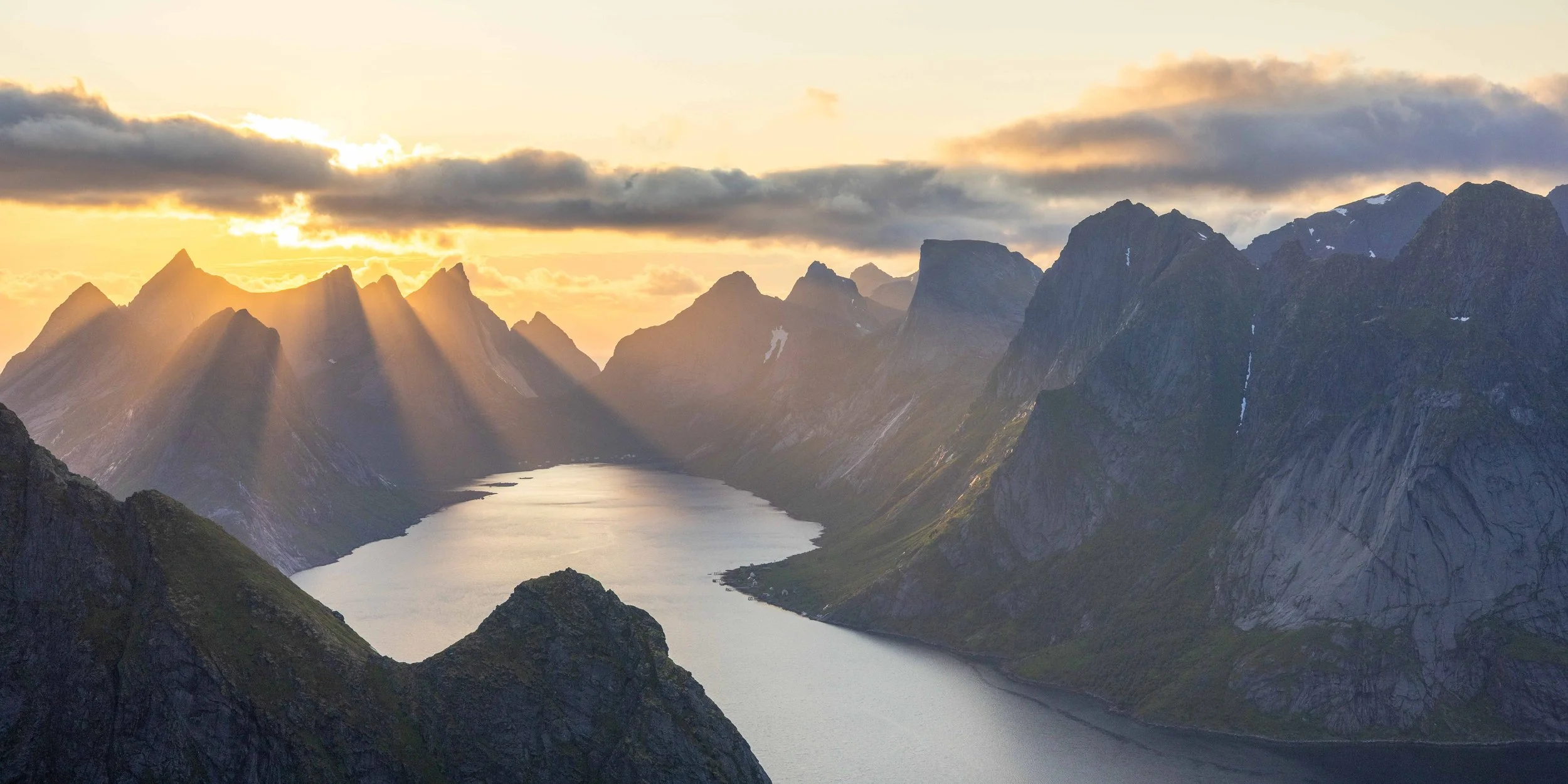Vista desde Reinebringen, Lofoten, con montañas dramáticas, rayos de luz dorada atravesando las nubes intensas y reflejos sobre el agua.