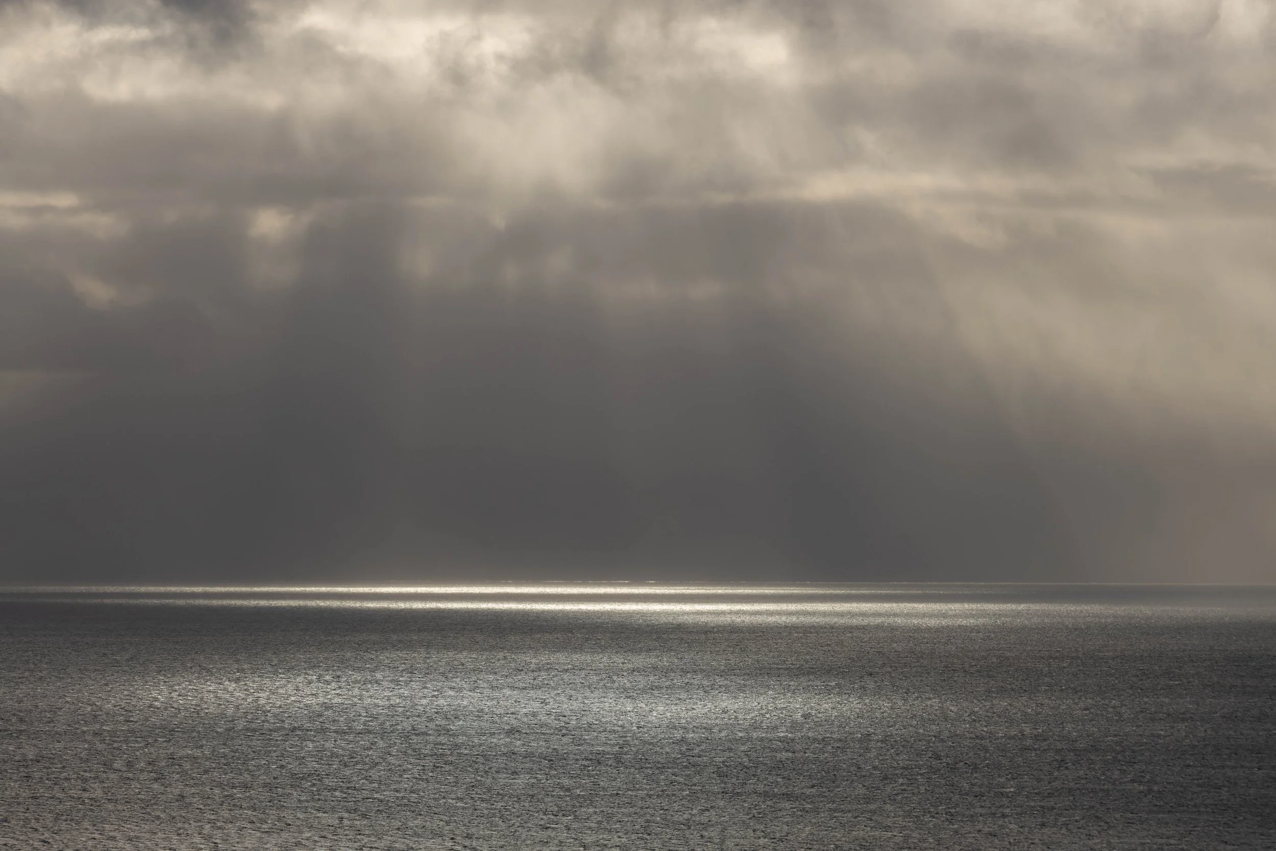Fotografía atmosférica del océano con rayos de sol atravesando nubes dramáticas y reflejos plateados sobre la superficie del mar.