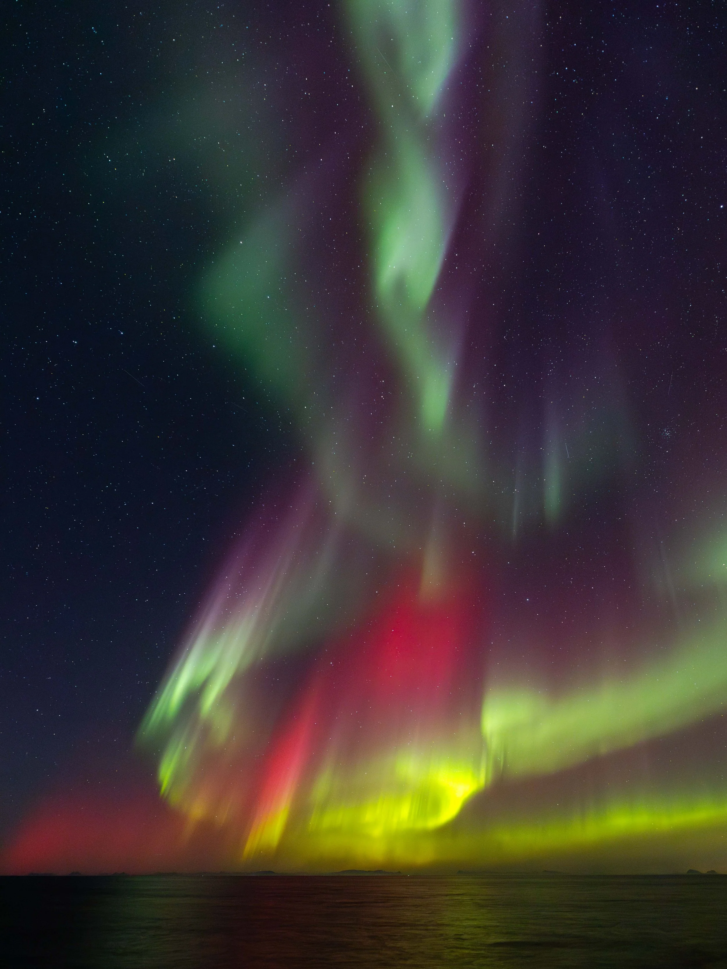 Imagen vertical de aurora boreal sobre el océano Ártico, con cortinas verdes, rojas, amarillas y violetas reflejándose suavemente sobre el mar en Noruega.