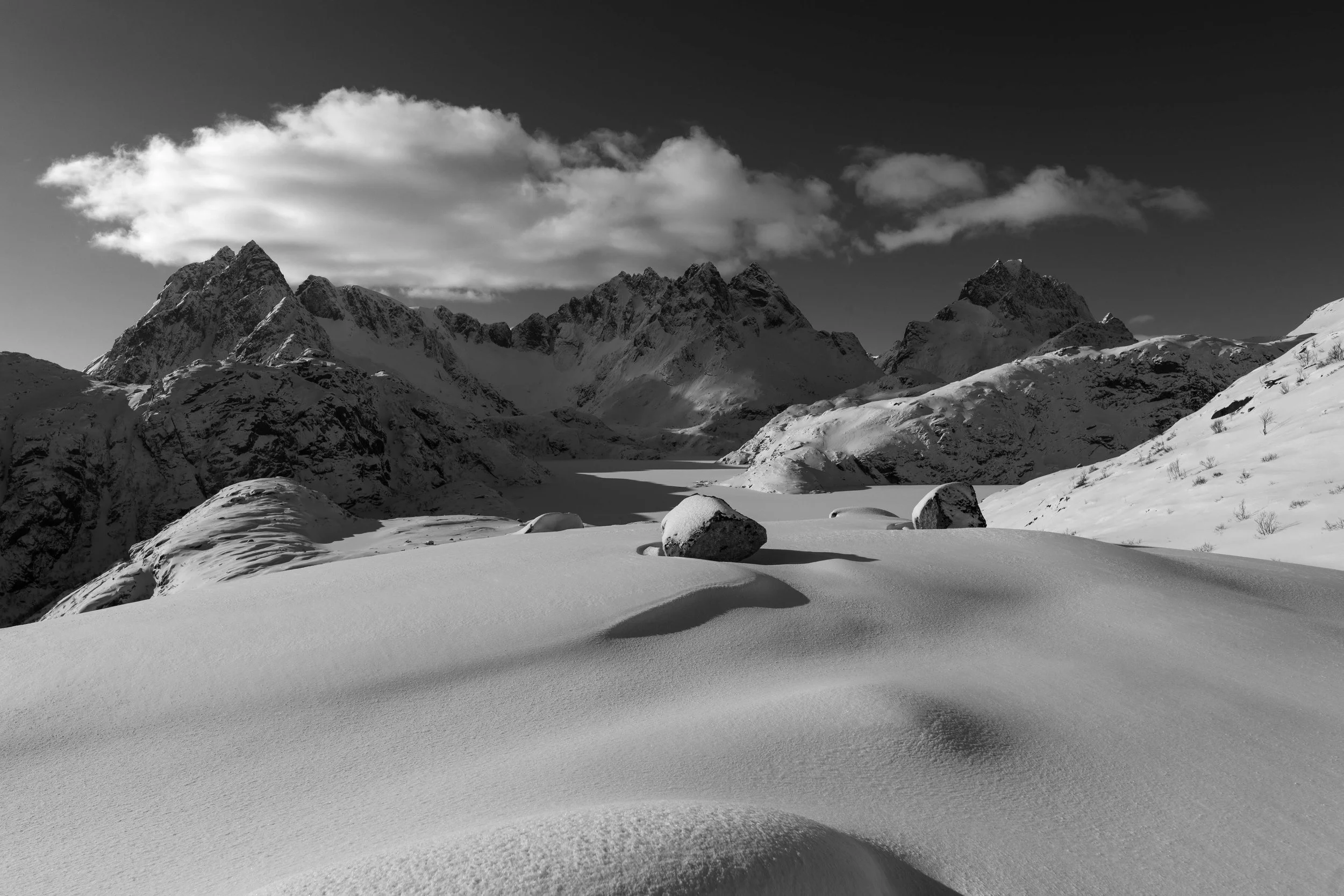 Fotografía artística en blanco y negro de un paisaje alpino cubierto de nieve, con montañas dramáticas, sombras profundas y textura suave en la nieve. Lofoten
