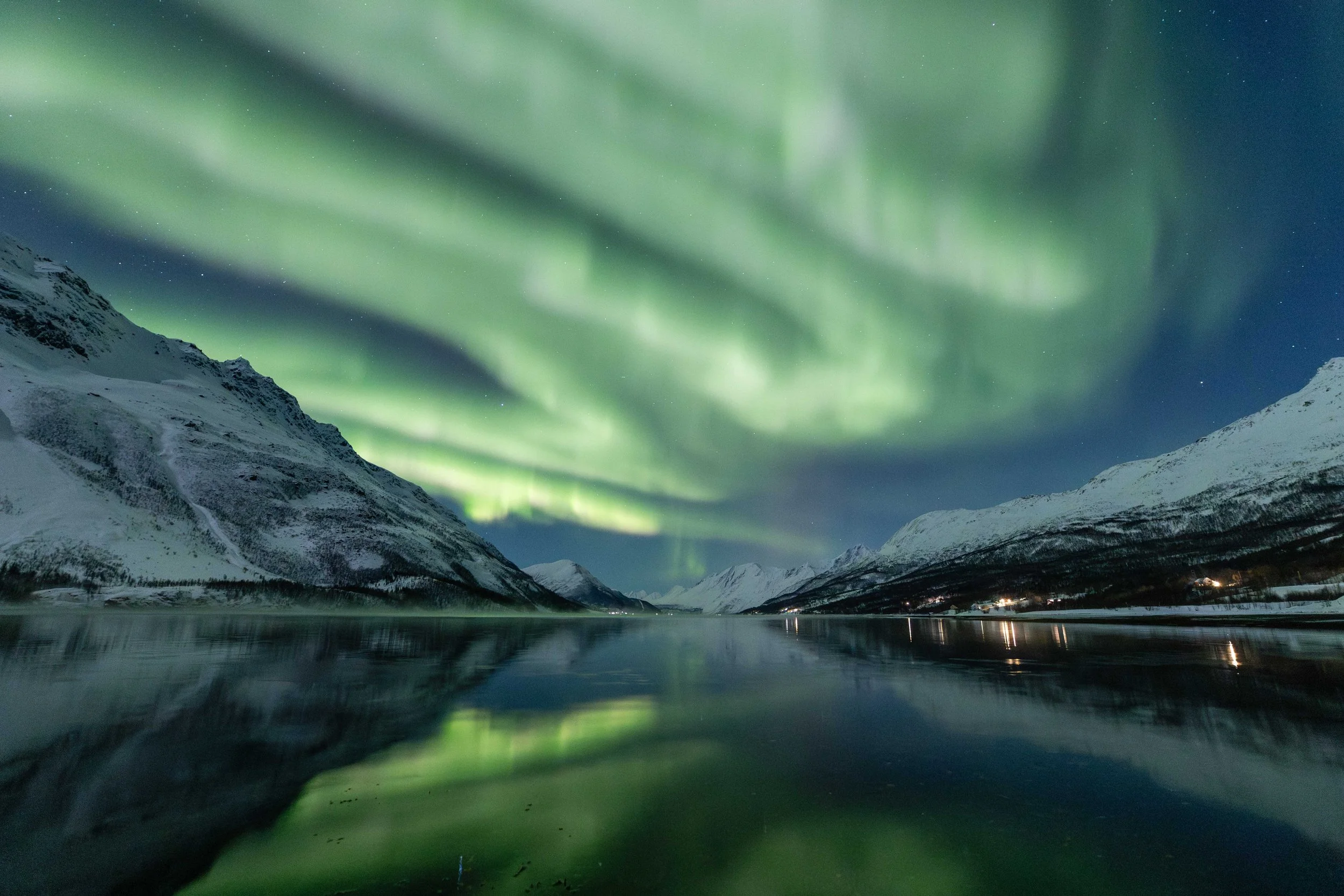 Imagen de aurora boreal cubriendo todo el cielo y reflejandose sobre un fiordo noruego, con montañas cubiertas de nieve.
