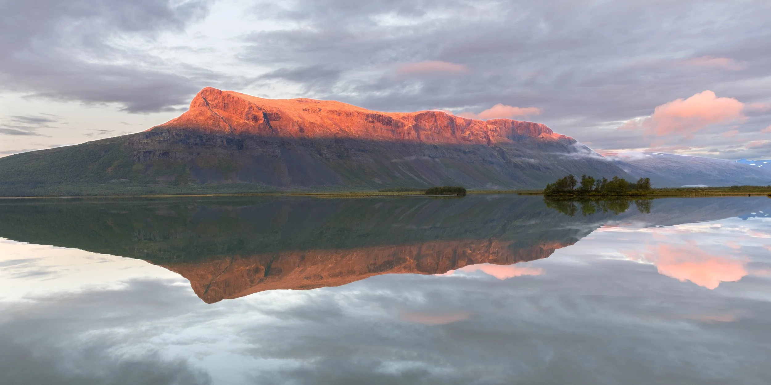 Fotografía panorámica de una montaña ártica luminada por el atardecer, con reflejo simétrico sobre aguas tranquilas y nubes suaves en el cielo. Sarek, Suecia