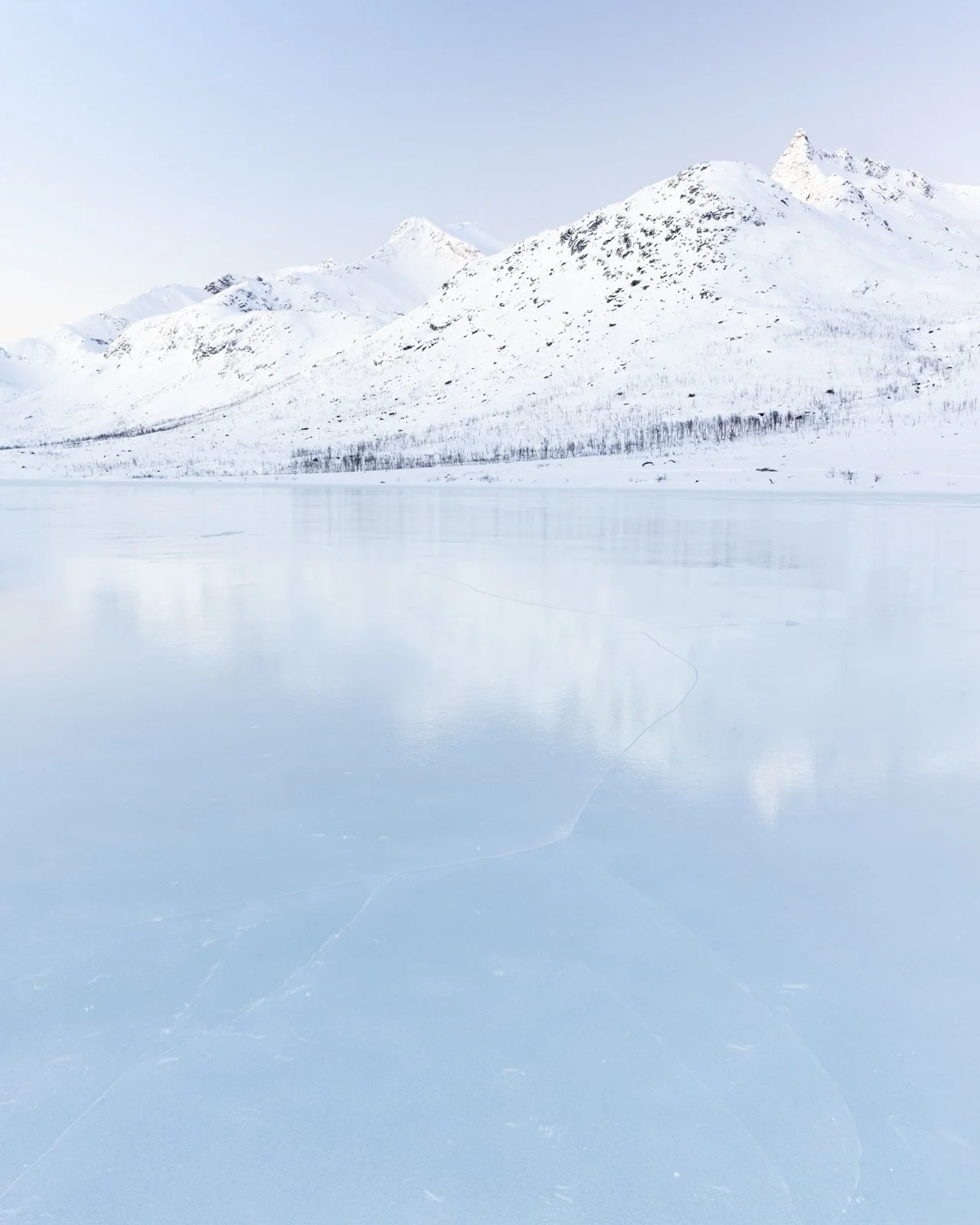 Fotografía minimalista de un paisaje invernal en Tromsø, Noruega, con un lago congelado, montañas nevadas y tonos fríos que transmiten calma y silencio ártico.
