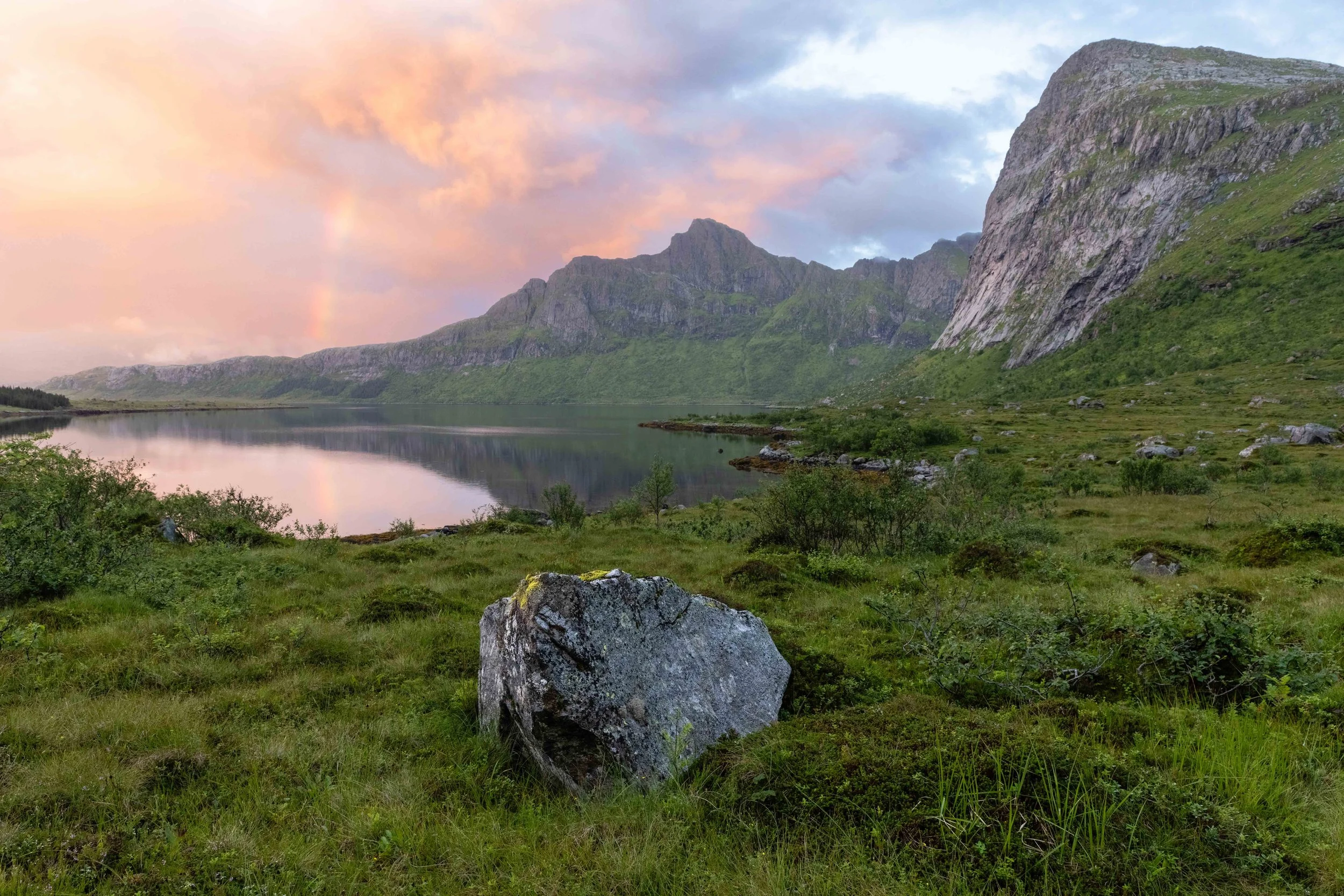 Paisaje de Lofoten en verano con fiordo en calma, montañas verdes, rocas, vegetación ártica y arcoiris suave al atardecer reflejado en el agua.